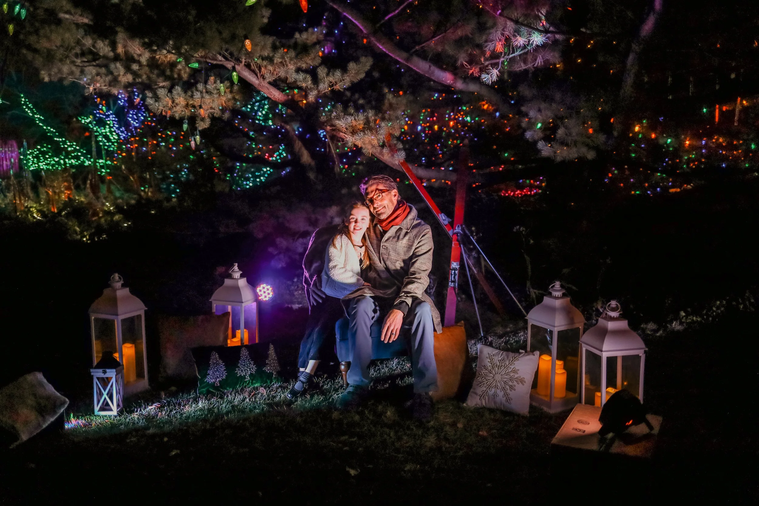 A man and a young girl sitting together under a decorated Christmas tree at night. The tree is adorned with multicolored lights, and the scene is decorated with lanterns, candles, and holiday-themed pillows.