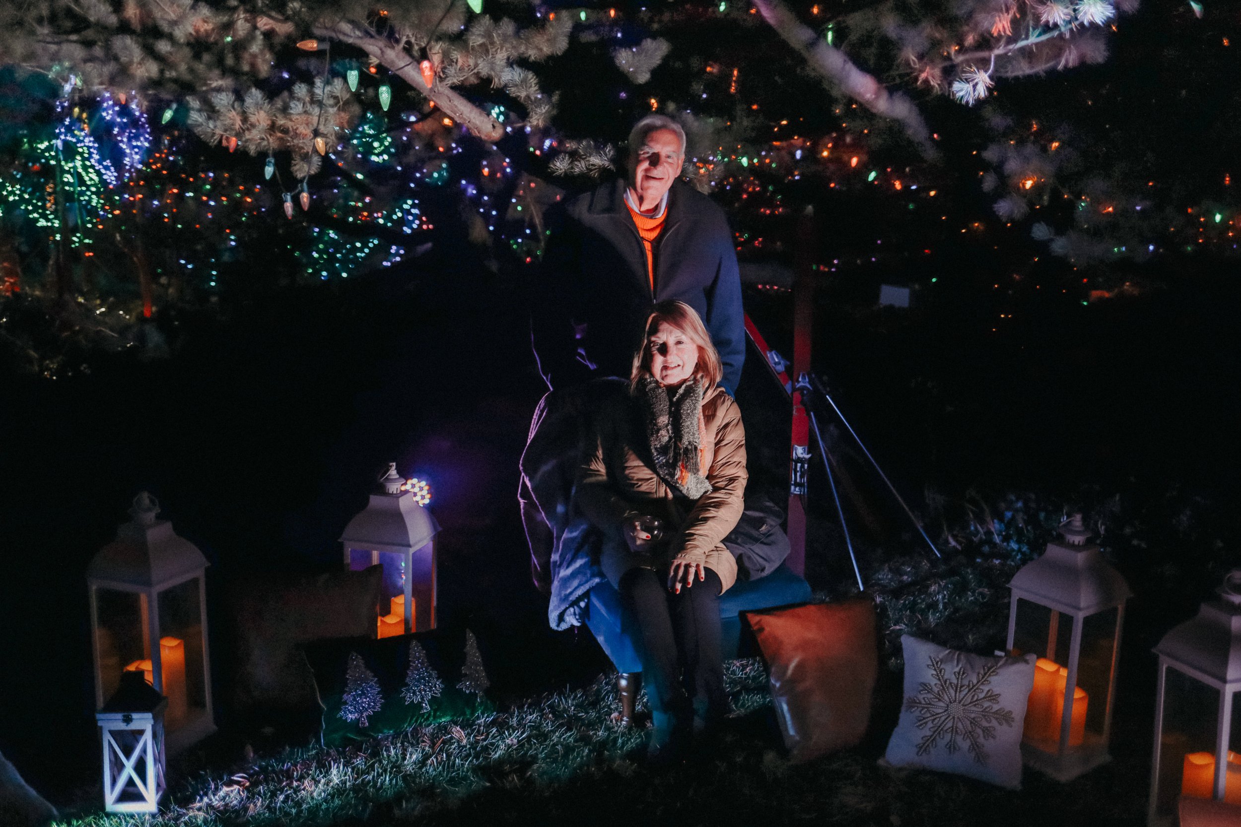 A man and woman are sitting in a decorative outdoor setting under a large, decorated Christmas tree at night. The area is illuminated with colorful lights, lanterns, and candles, with Christmas pillows nearby.
