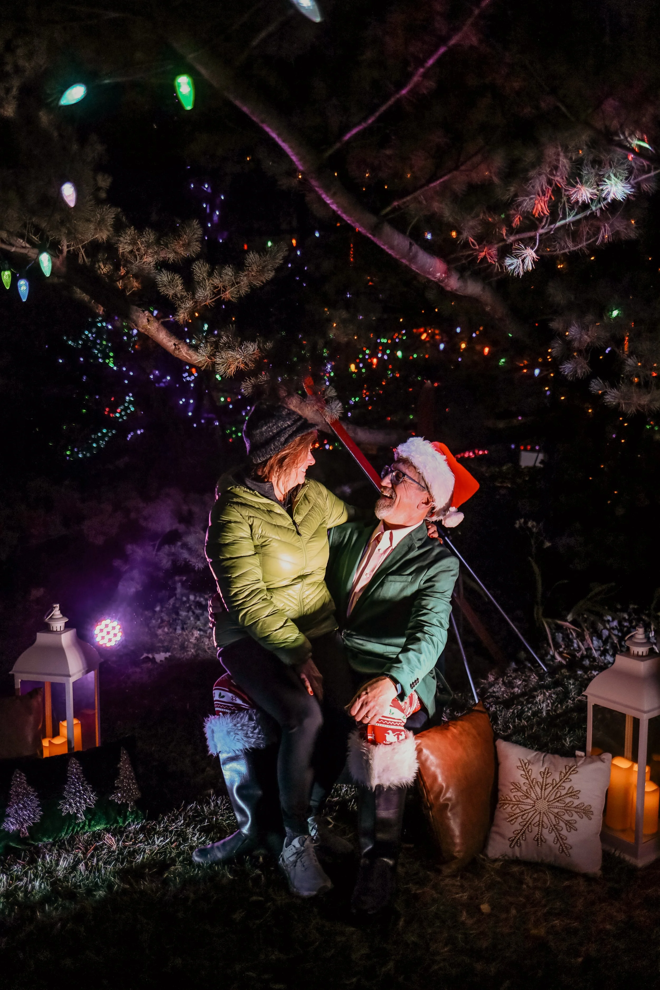 A couple sitting on a swing under a decorated Christmas tree at night, wearing holiday attire including a Santa hat and festive clothing, surrounded by candles and holiday decorations.