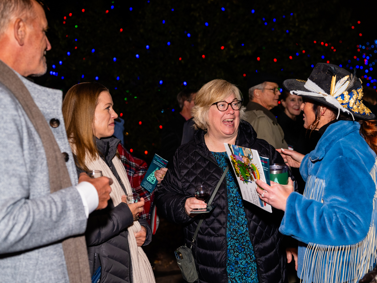 Group of people enjoying a festive outdoor event at night with colorful Christmas lights in the background. Woman in a black jacket holding a glass of wine appears to be laughing and talking to a woman dressed in a blue coat and a decorative wide-bri