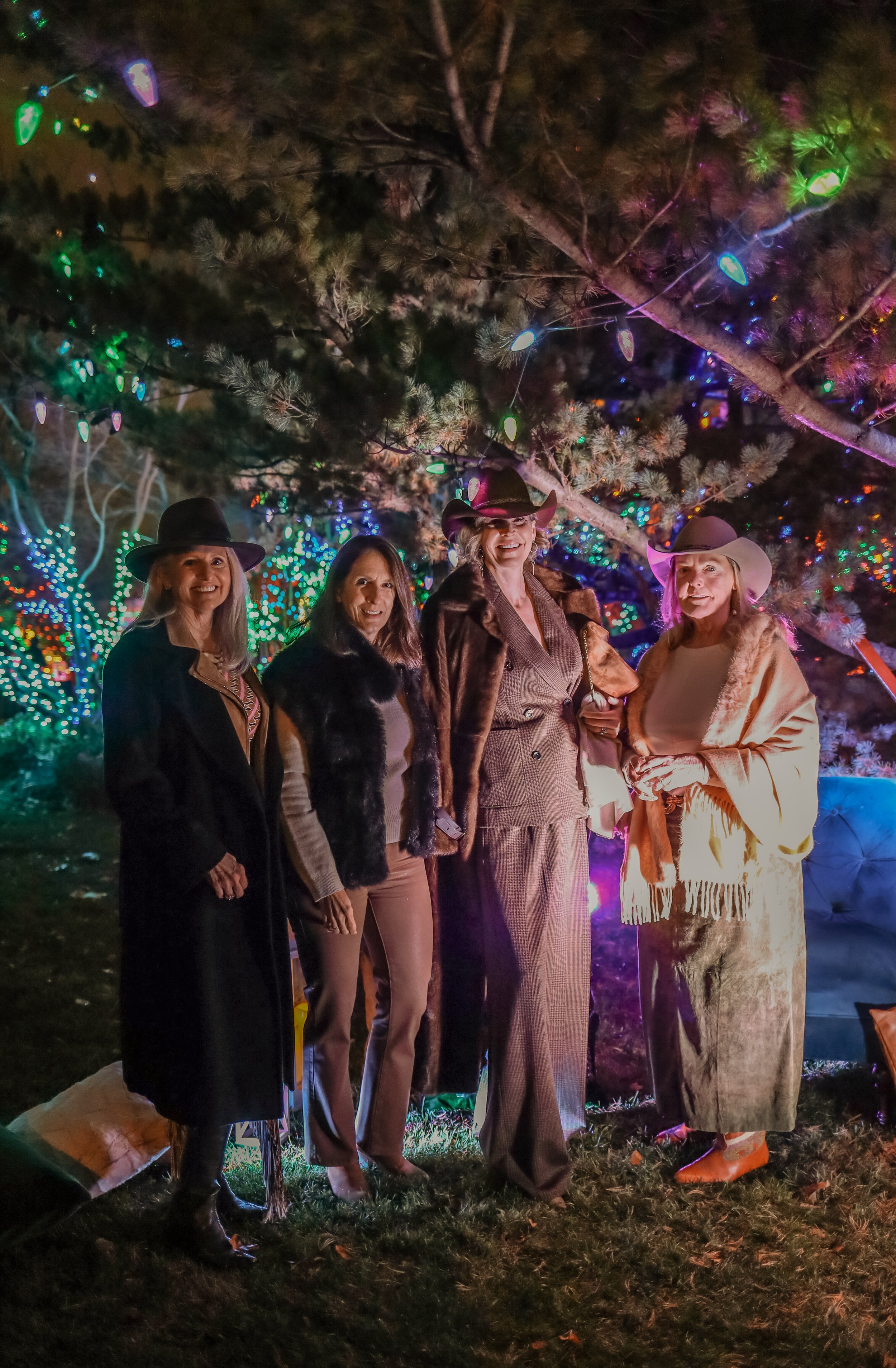 Four women dressed in Western-style clothing, standing outdoors at night under a decorated tree with colorful lights.