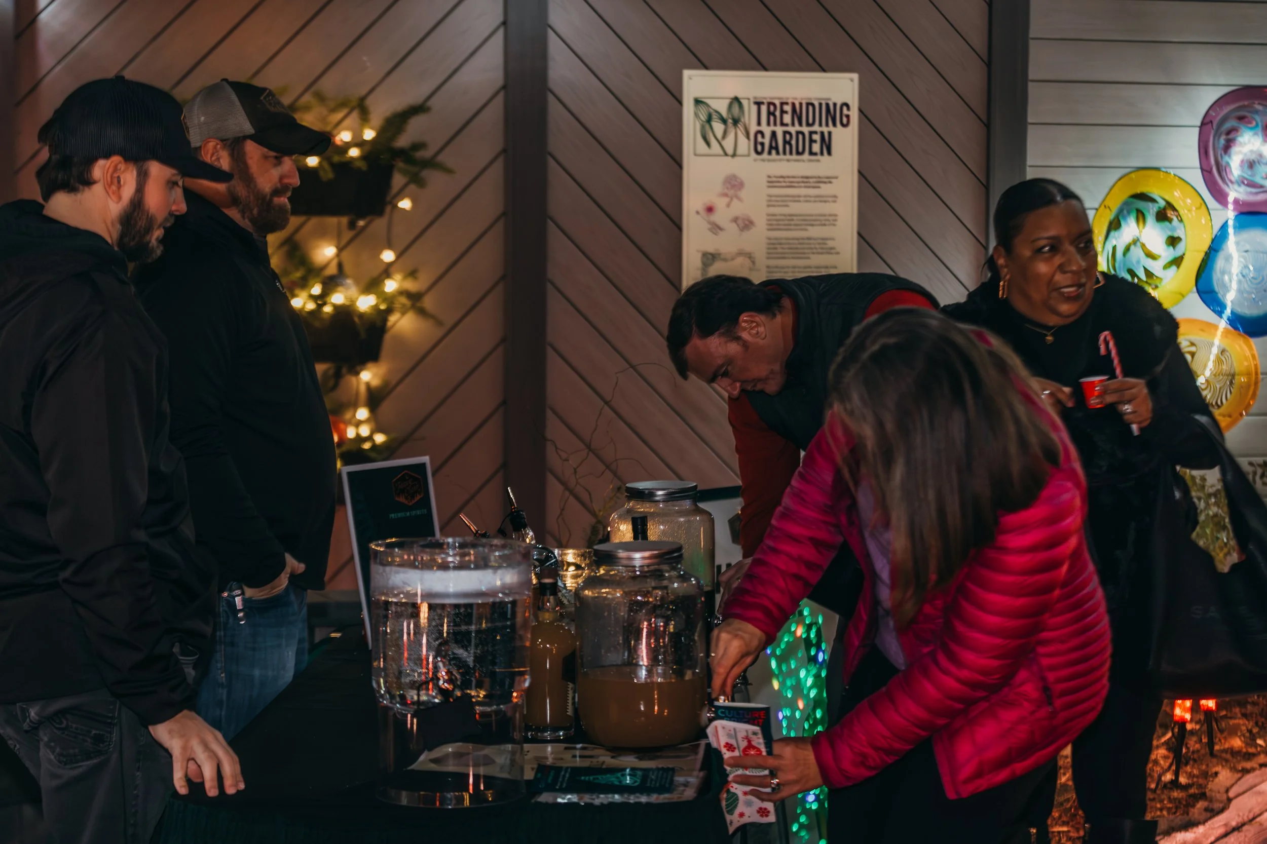 People at a booth with jars of liquids on a table at an indoor event, festive balloons, and a sign that says 'Trending Garden.'