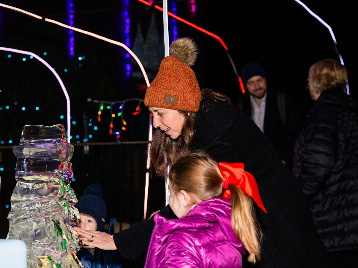 People gathered around an ice sculpture at a nighttime outdoor event with colorful string lights in the background.