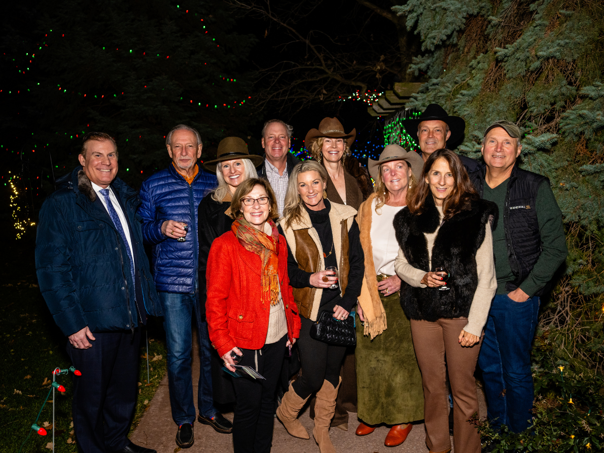 A group of people standing outdoors at night, smiling and holding glasses of wine, with Christmas lights and trees in the background.