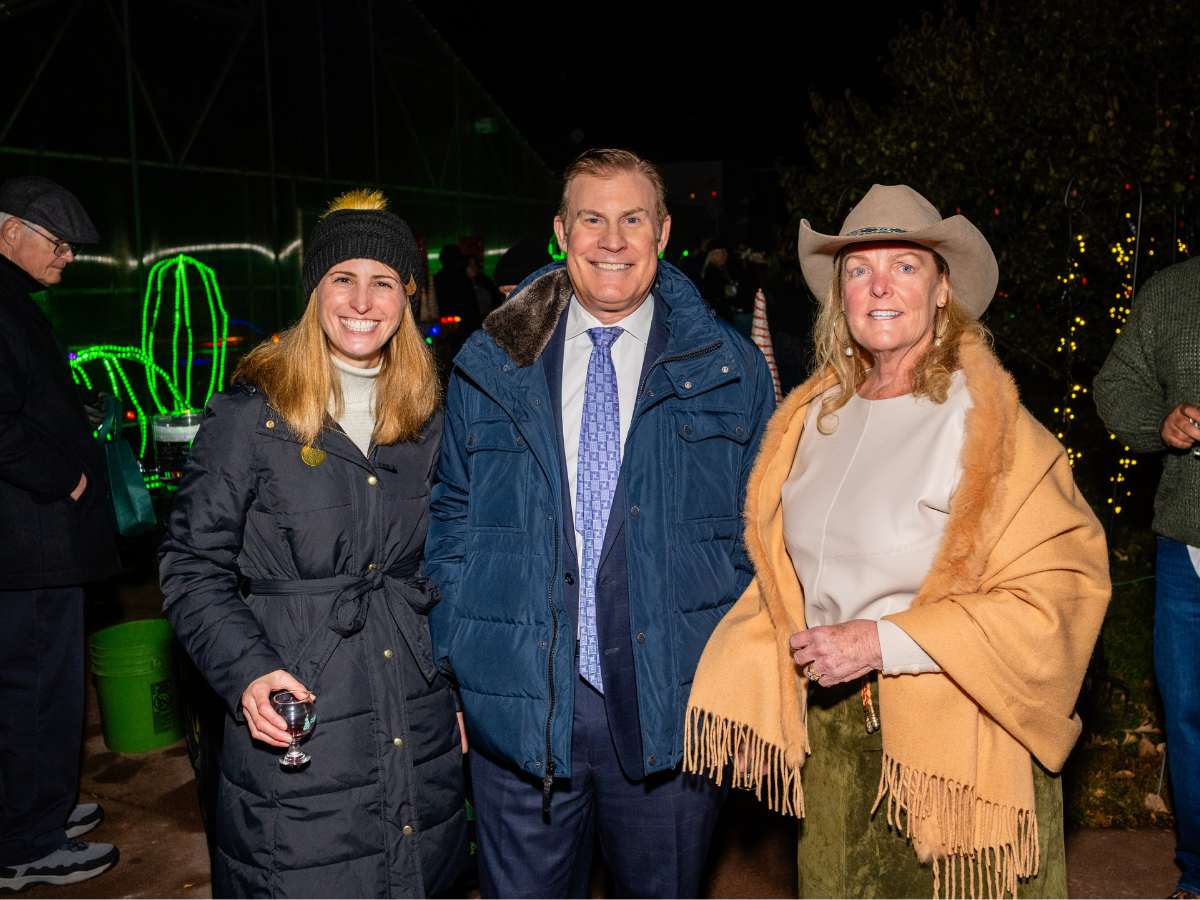 Three people standing together outdoors at night, smiling for the camera. The woman on the left is wearing a dark coat, a gray hat with a pom-pom, and holding a glass of wine. The man in the middle is in a blue jacket and a tie, smiling brightly. The