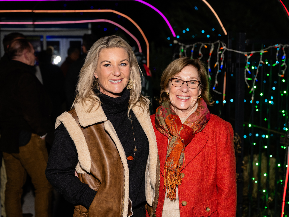 Two women smiling at an event with colorful lights in the background, one in a tan and black jacket, and the other in a red coat and scarf.