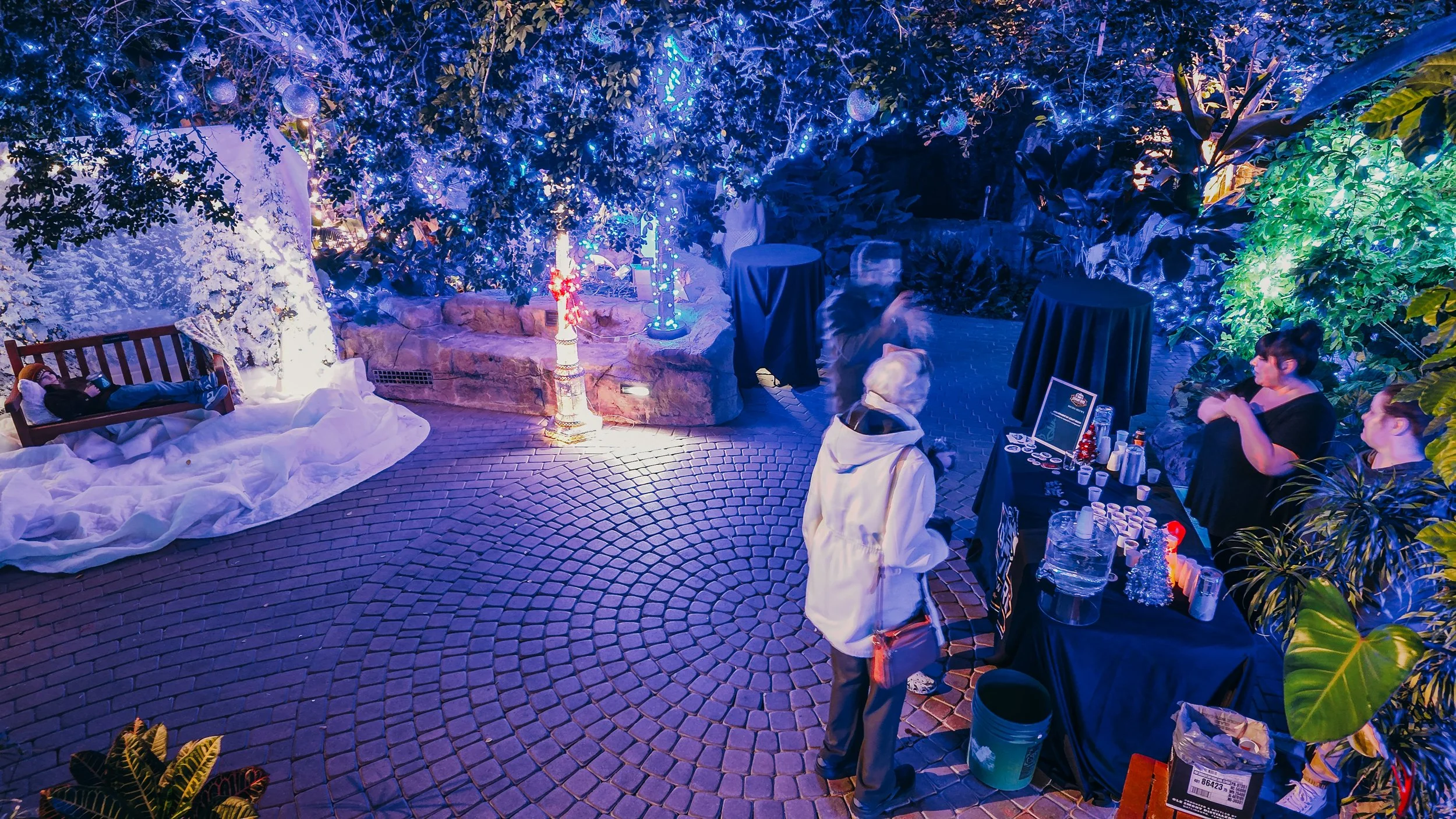Outdoor evening scene with a person lying on a wooden bench with white blankets, surrounded by illuminated blue and purple lights, trees, and plants. Several people are gathered around a black table with drinks and snacks. The area is decorated with 
