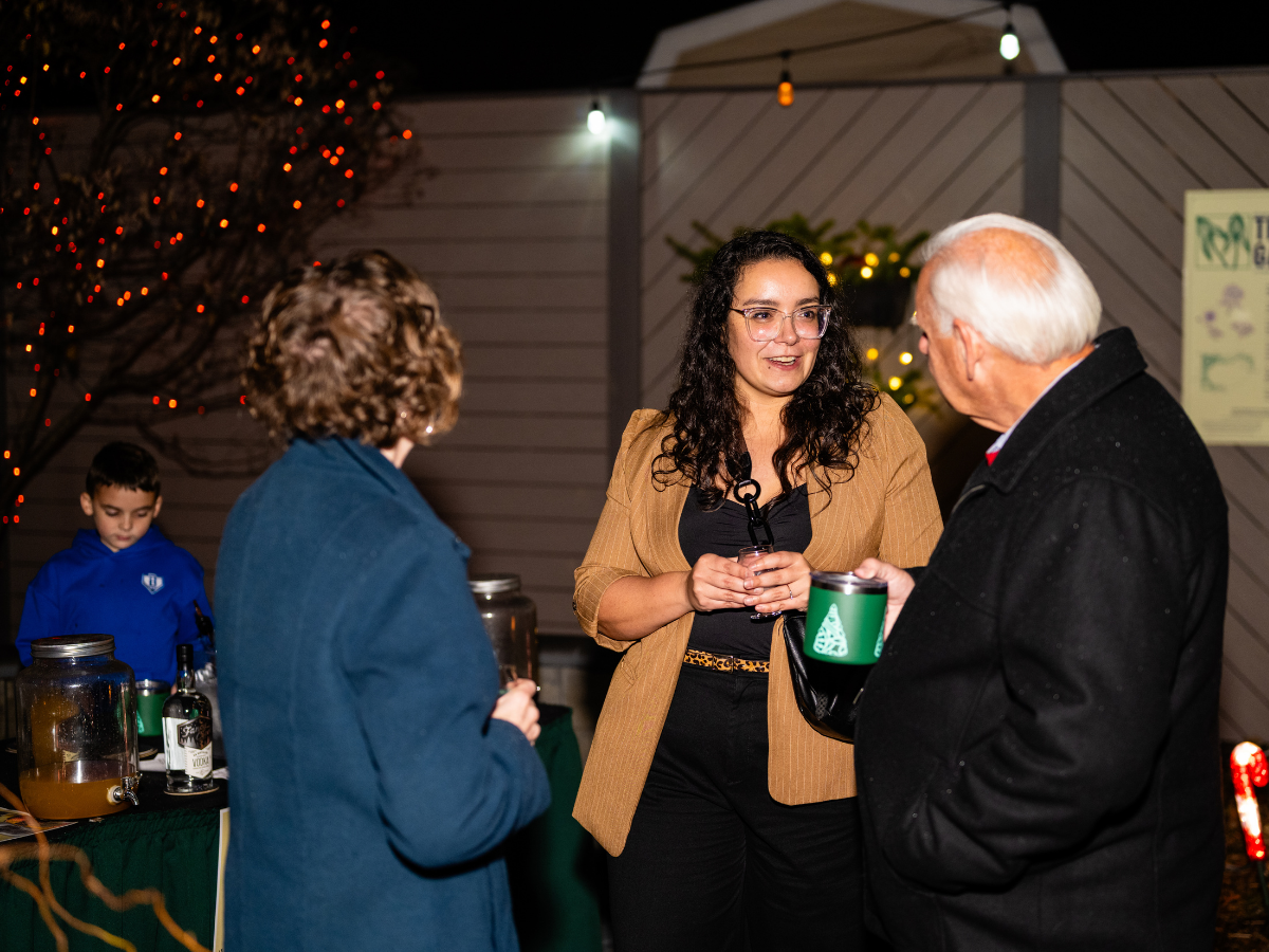 Three people engaging in conversation at an outdoor event with Christmas decorations, including a tree with red lights, in the background. A vendor table with drinks and bottles is visible in the background, along with a young boy in a blue hoodie.