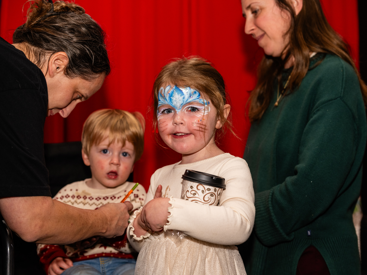 A young girl with face paint of a blue whale holding a coffee cup, standing among two women and a boy, with a red curtain in the background.