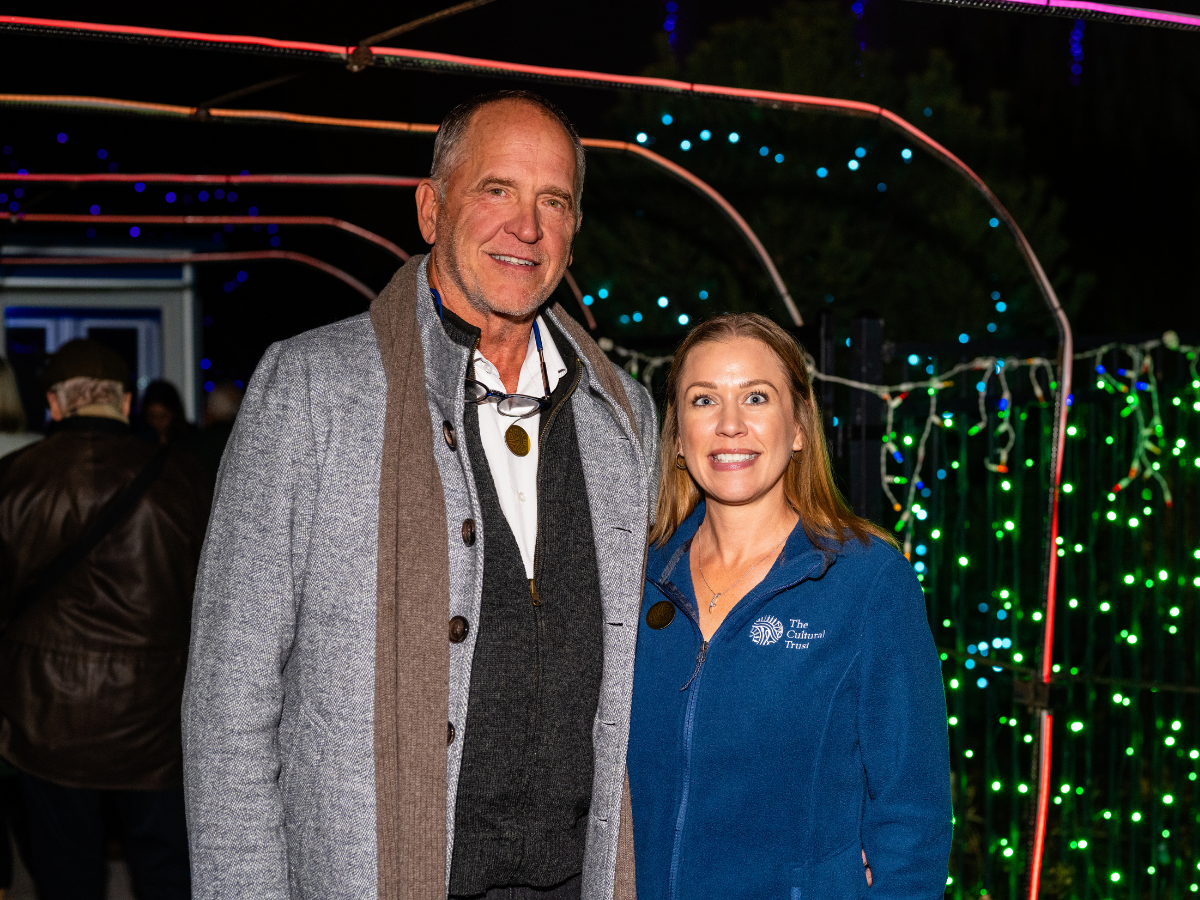 Two people standing outdoors at night, smiling, with colorful string lights in the background.