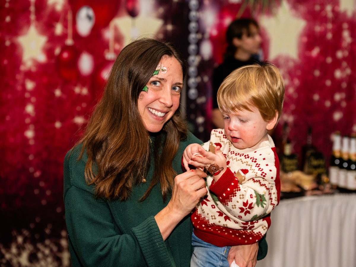 A woman with long brown hair wears a green sweater and smiles while holding a young boy with blond hair, who wears a festive Christmas sweater with red and white patterns. They are in front of a decorated red background with Christmas ornaments and l