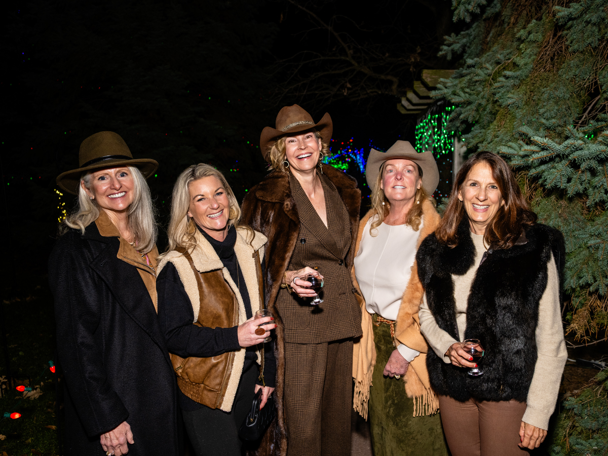 Five women dressed in Western and fall attire, holding wine glasses, standing outdoors at night with Christmas lights and evergreen trees in the background.