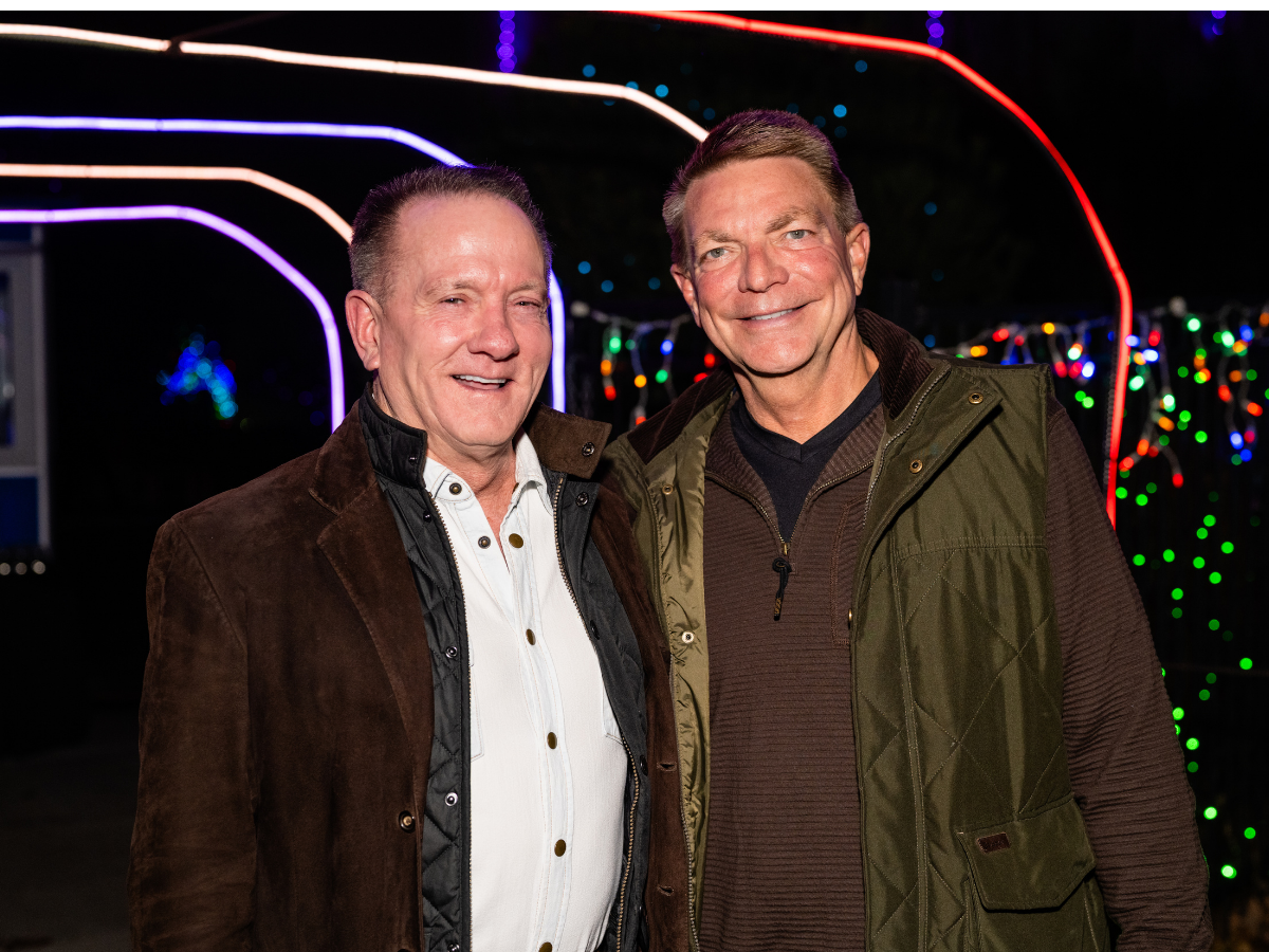 Two smiling middle-aged men standing close together outdoors at night, with colorful string lights in the background.