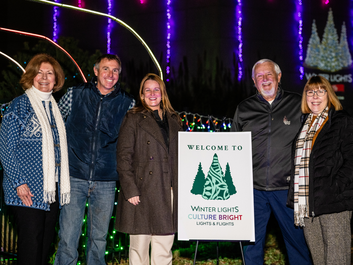 A group of six people standing outdoors at night, smiling, in front of colorful Christmas and winter light decorations. There is a sign that reads "Welcome to Winter Lights. Culture Bright. Lights & Flights."