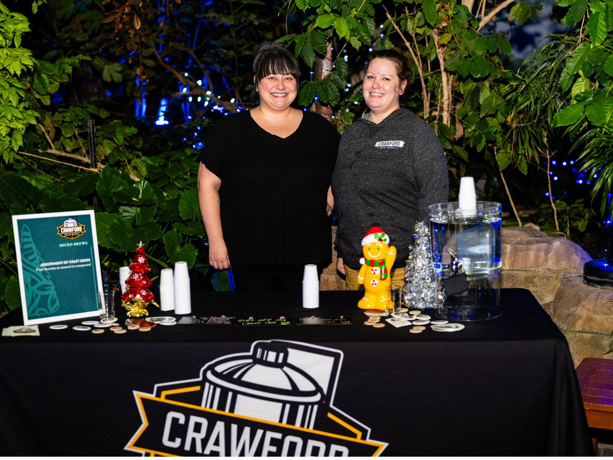 Two women standing behind a black table at a festive event, with greenery and string lights in the background. The table has a sign for Crawford Micro-Brews, a gingerbread decoration, a water jar, and small paper cups. Both women are smiling; the wom