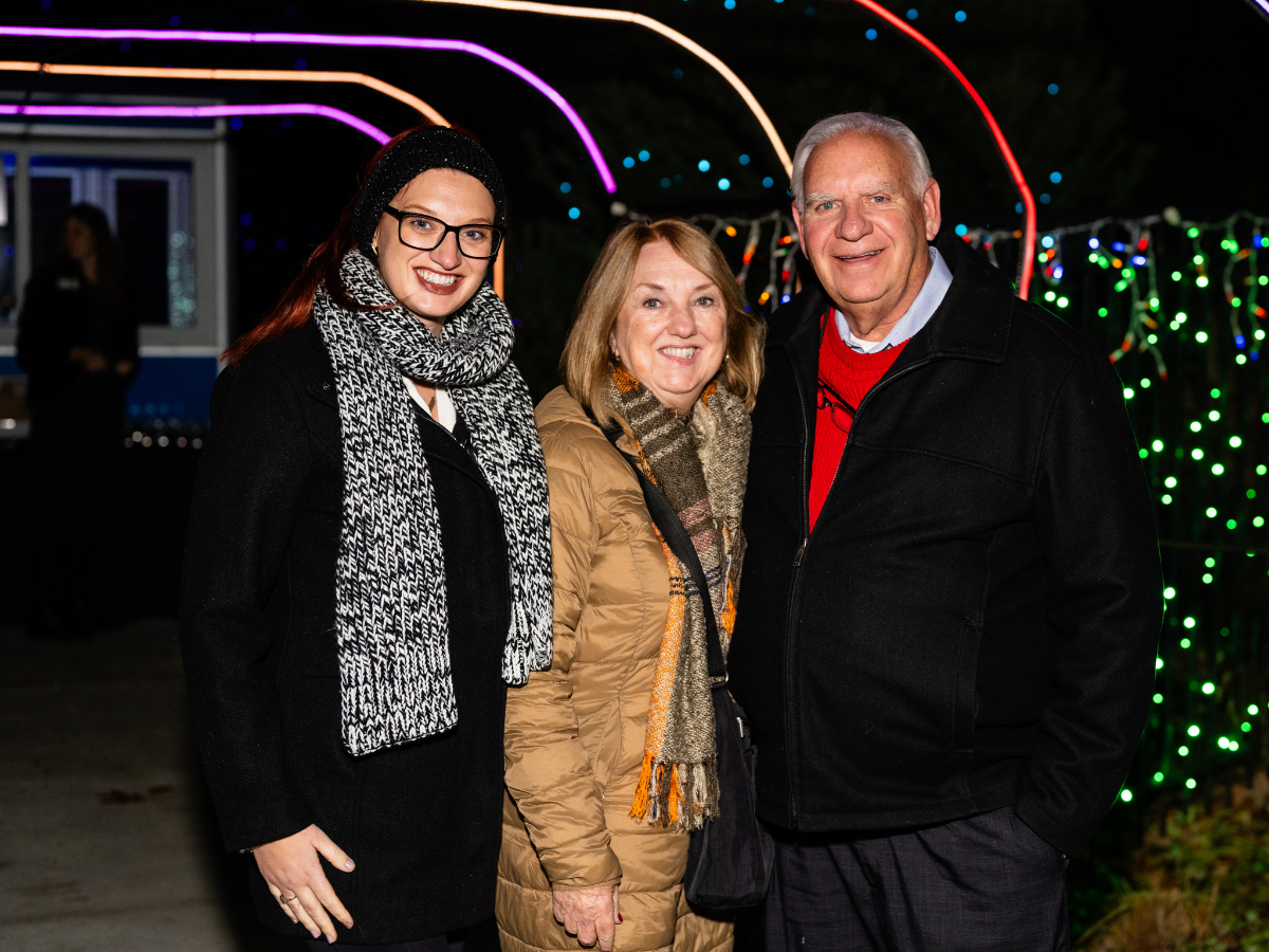 Three people standing outdoors at night, smiling with colorful holiday lights in the background.