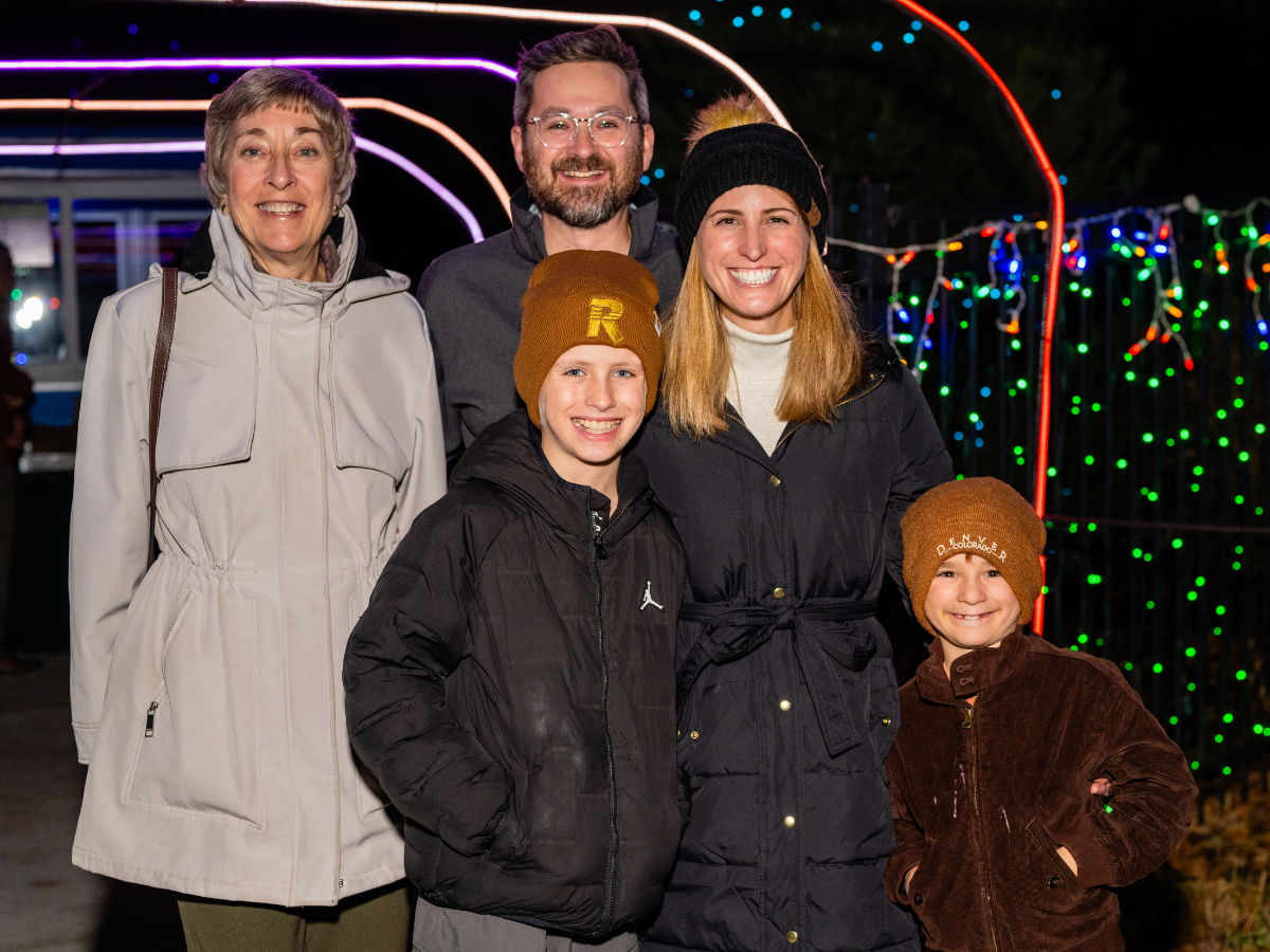 A smiling family of five, including two women, one man, and two boys, standing outdoors at night with colorful decorative lights behind them.