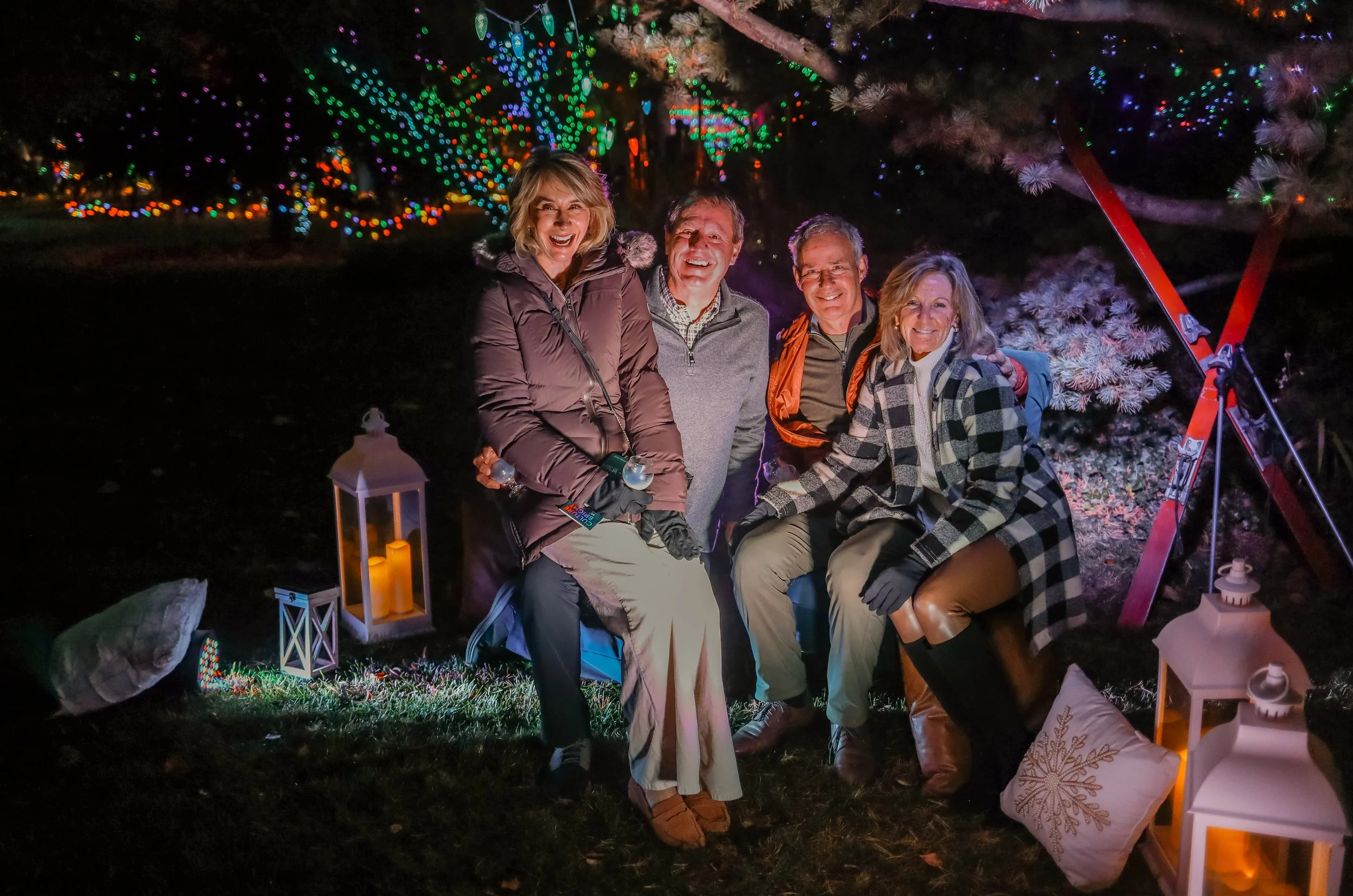 Four adults sitting outdoors at night during a holiday event, surrounded by colorful Christmas lights and festive decorations, smiling and enjoying the occasion.