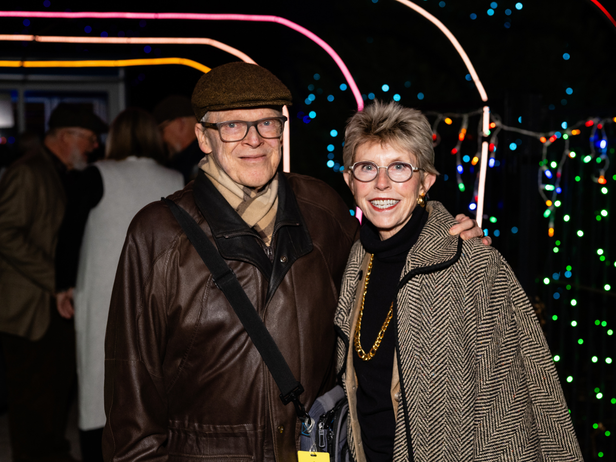 A man and woman smiling at an outdoor event with festive string lights in the background at night.