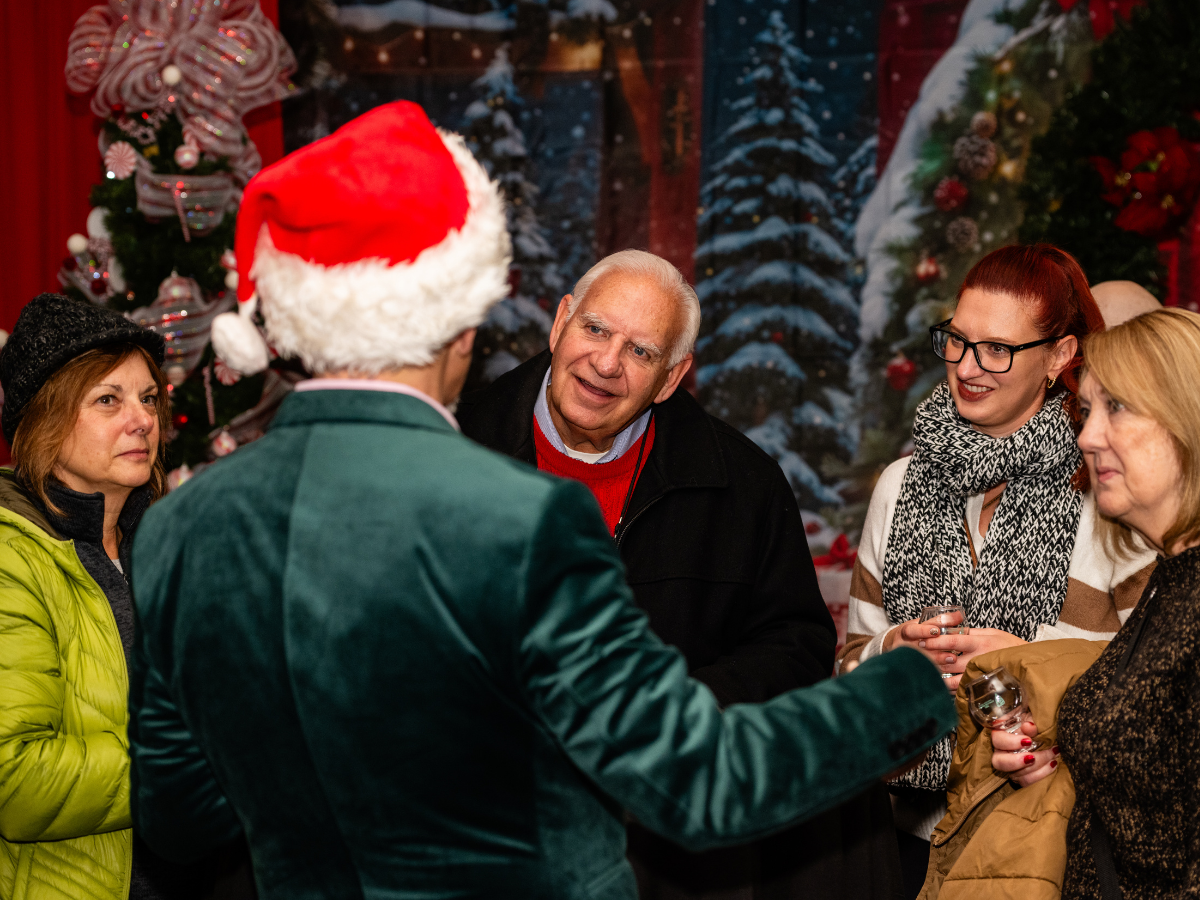 People at a Christmas party talking to a person dressed as Santa Claus, with festive holiday decorations and a Christmas tree in the background.