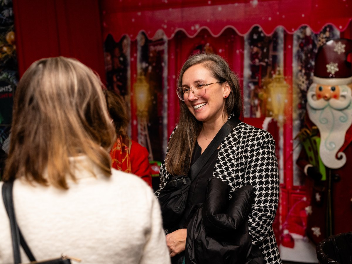A woman with glasses and long brown hair smiling and talking to another woman in front of a Christmas-themed background with decorations and a large Santa figure.