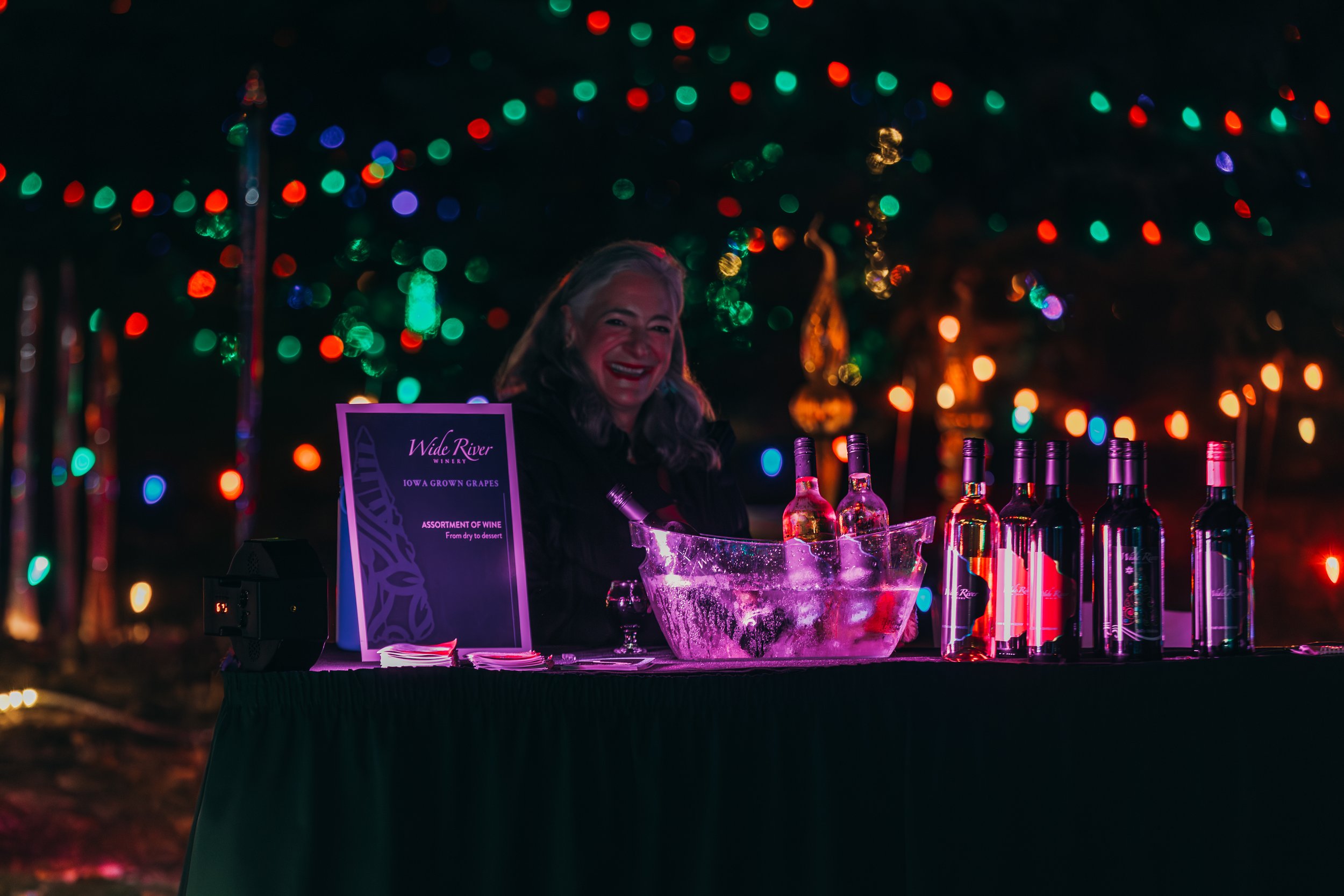 A woman smiling behind a table with bottles of wine and an ice bucket, festive lights in the background.