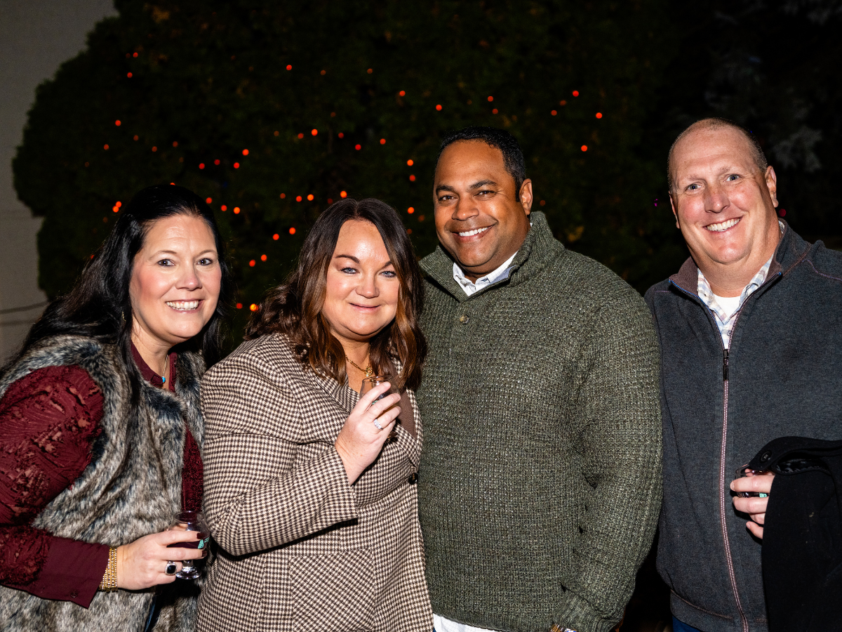 Group of four adults smiling at a nighttime outdoor event, with Christmas lights in the background.