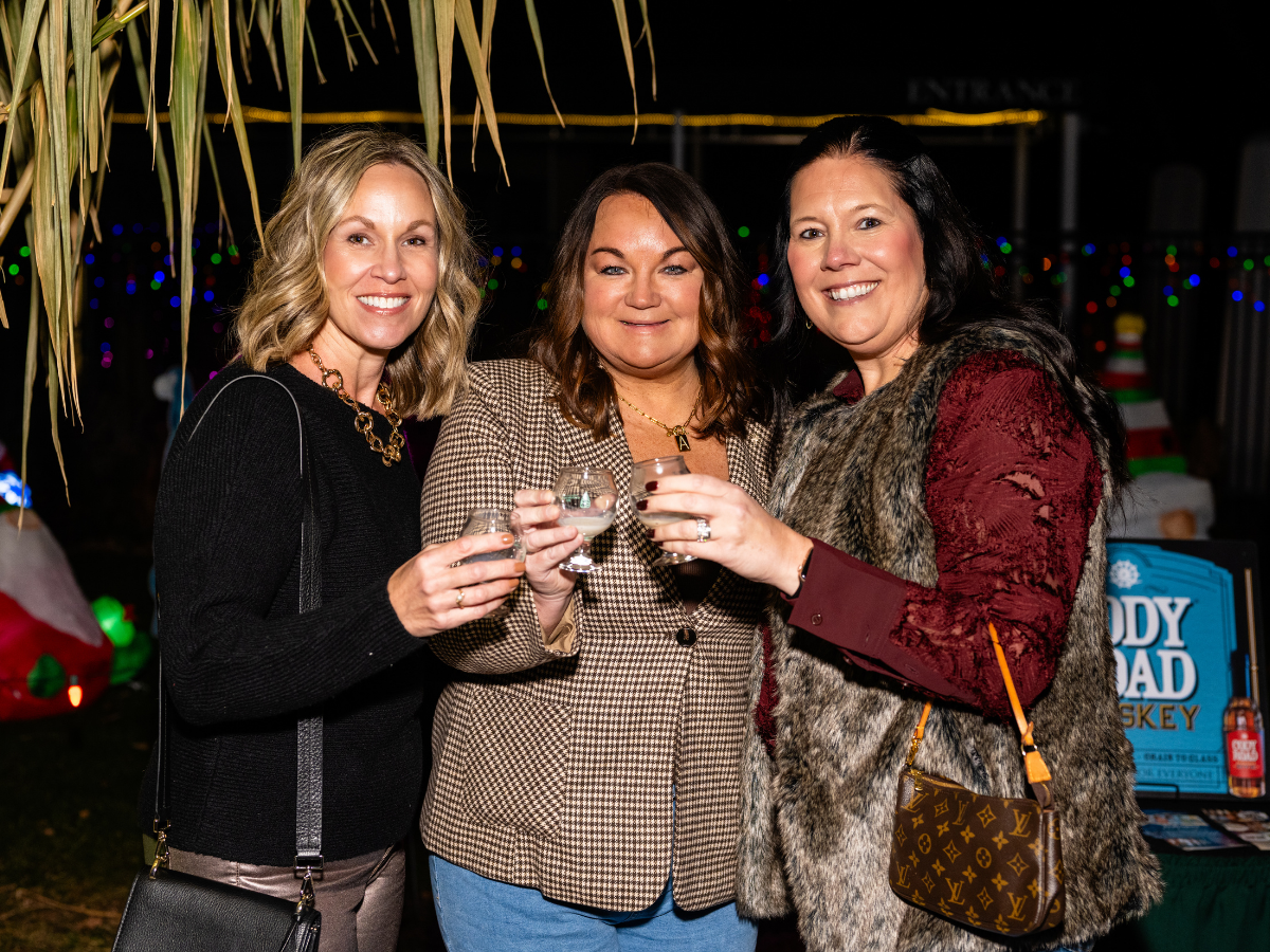 Three women smiling and holding drinks at an outdoor event during nighttime, with decorative lights in the background.
