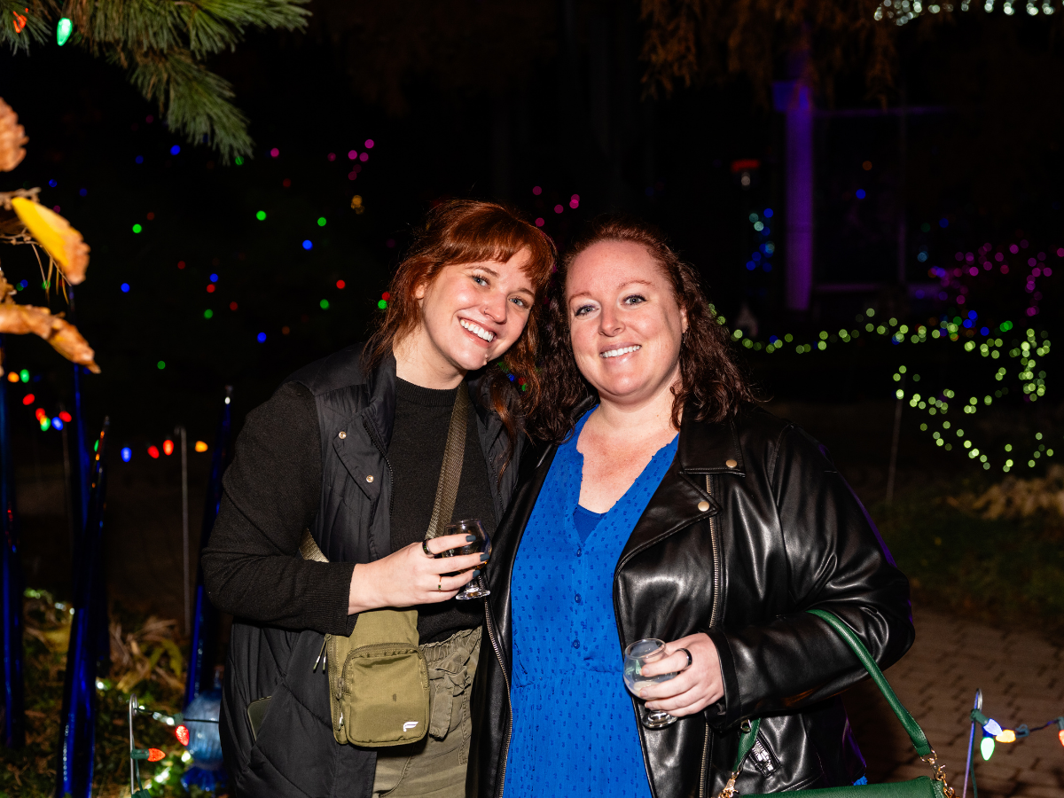 Two women smiling and holding drinks at nighttime outdoors, with colorful string lights and blurred decorations in the background.