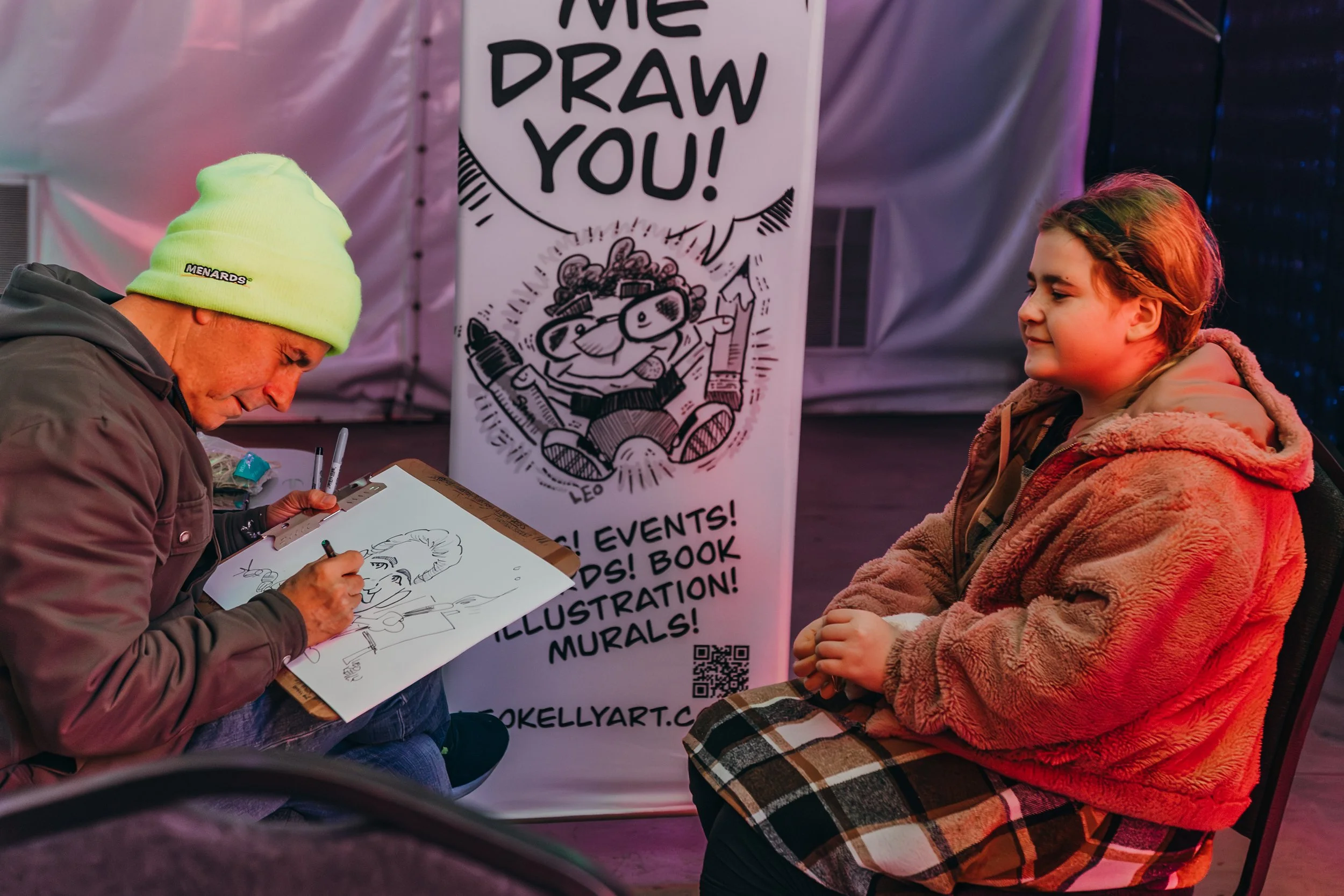 An artist drawing a caricature of a young girl at a festival or event. The artist is wearing a light green beanie and gray jacket, working on a sketchpad. Behind them, a banner reads 'Come Draw You!' with cartoon illustrations and event details.