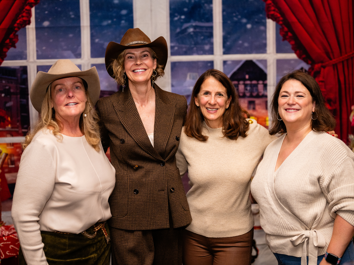 Four women smiling and standing together in front of a window with a snowy scene outside, dressed in casual and western-style clothing, two of them wearing cowboy hats.