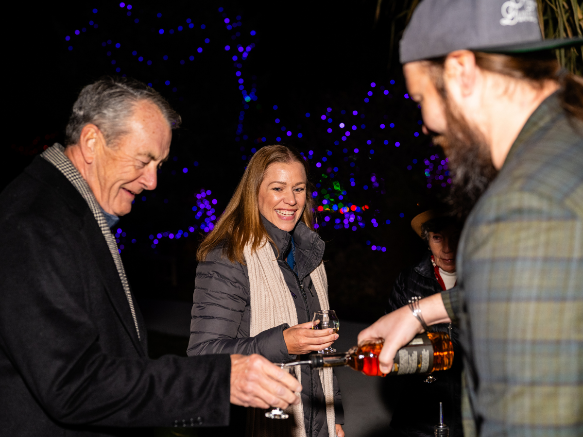 People celebrating outdoors at night, with colorful Christmas lights in the background, one man pouring a drink into a glass held by a woman smiling.