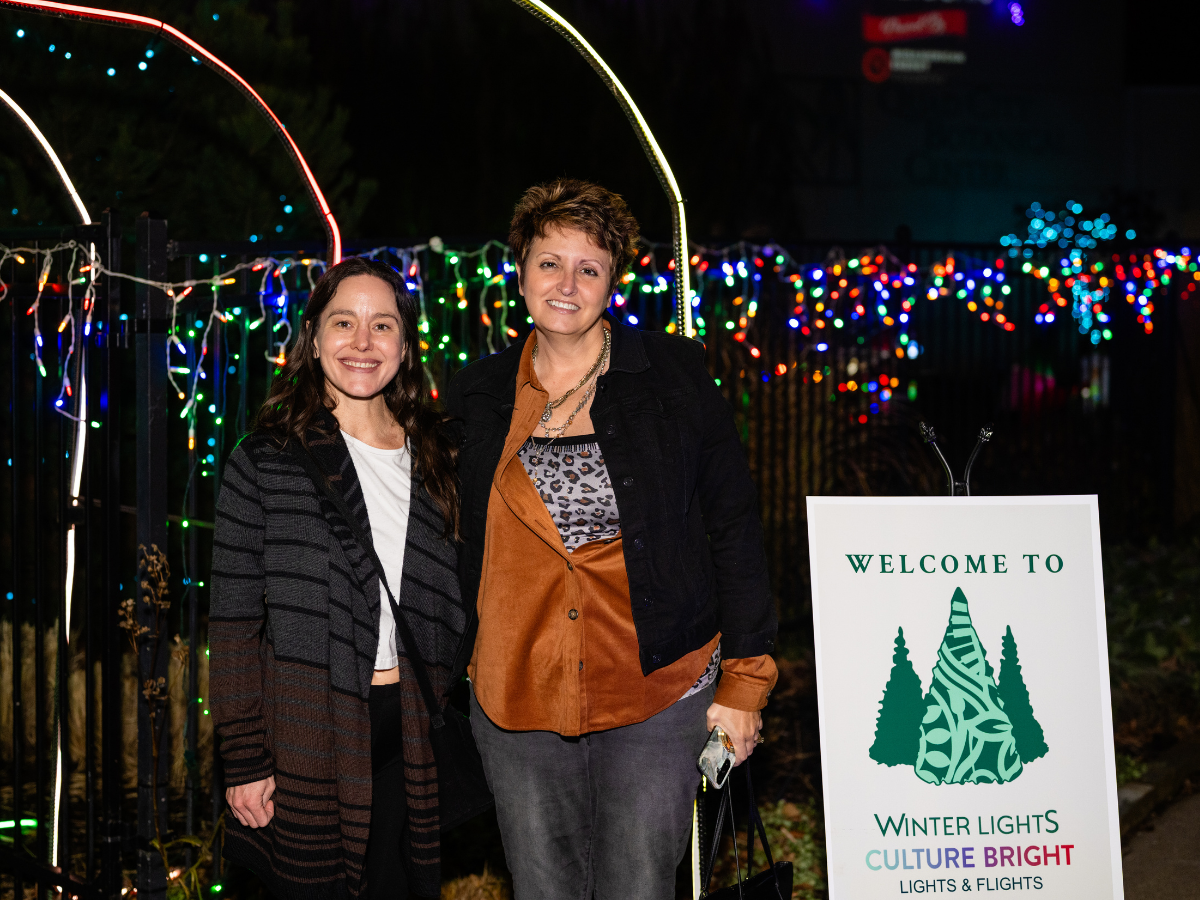 Two women standing outdoors at night in front of a fence decorated with colorful Christmas lights. One woman is young with long dark hair, wearing a striped cardigan. The other woman is older with short hair, wearing a black jacket over a patterned s