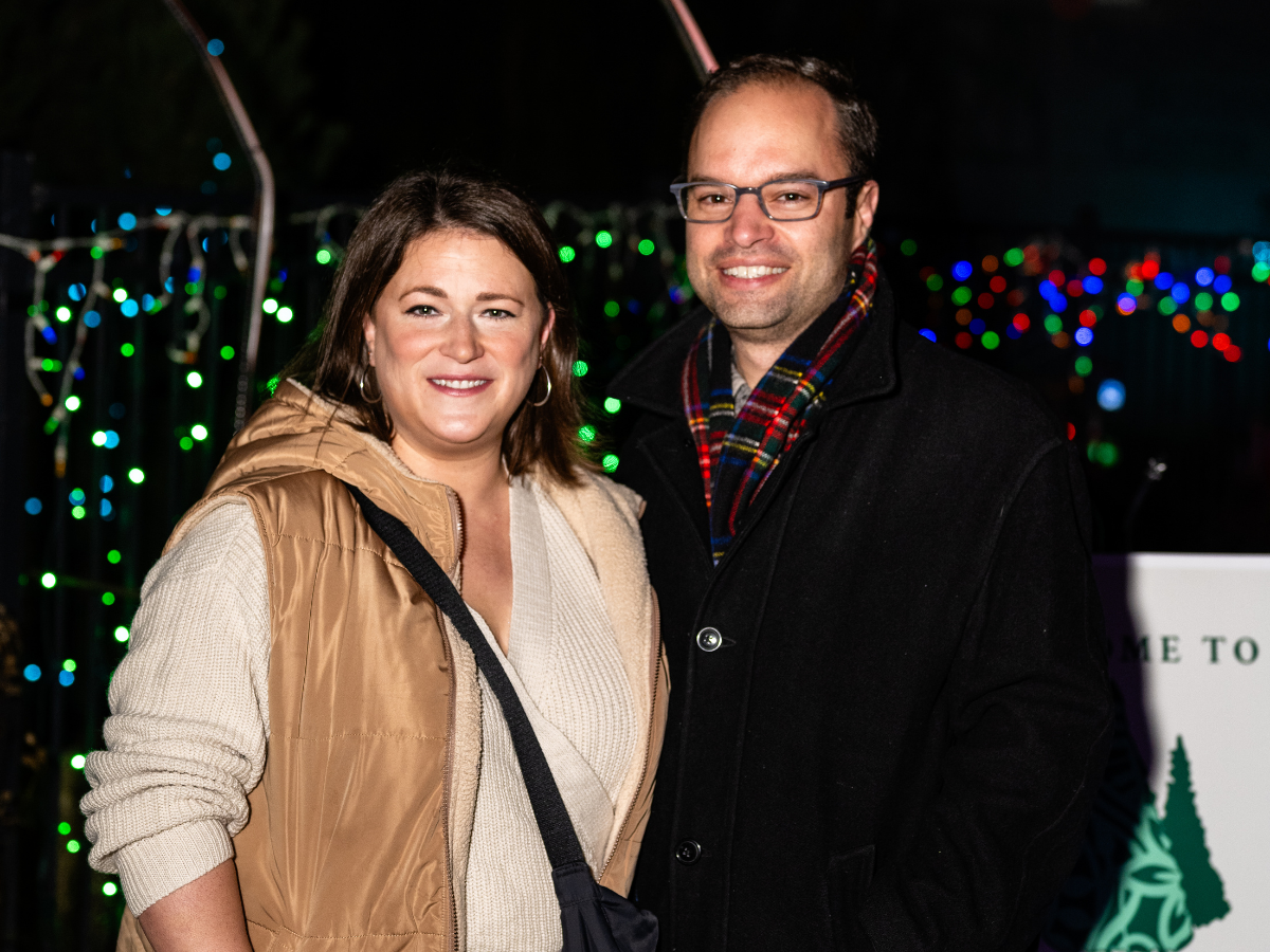 A smiling woman and man standing close together outdoors at night, with colorful string lights in the background, dressed warmly.