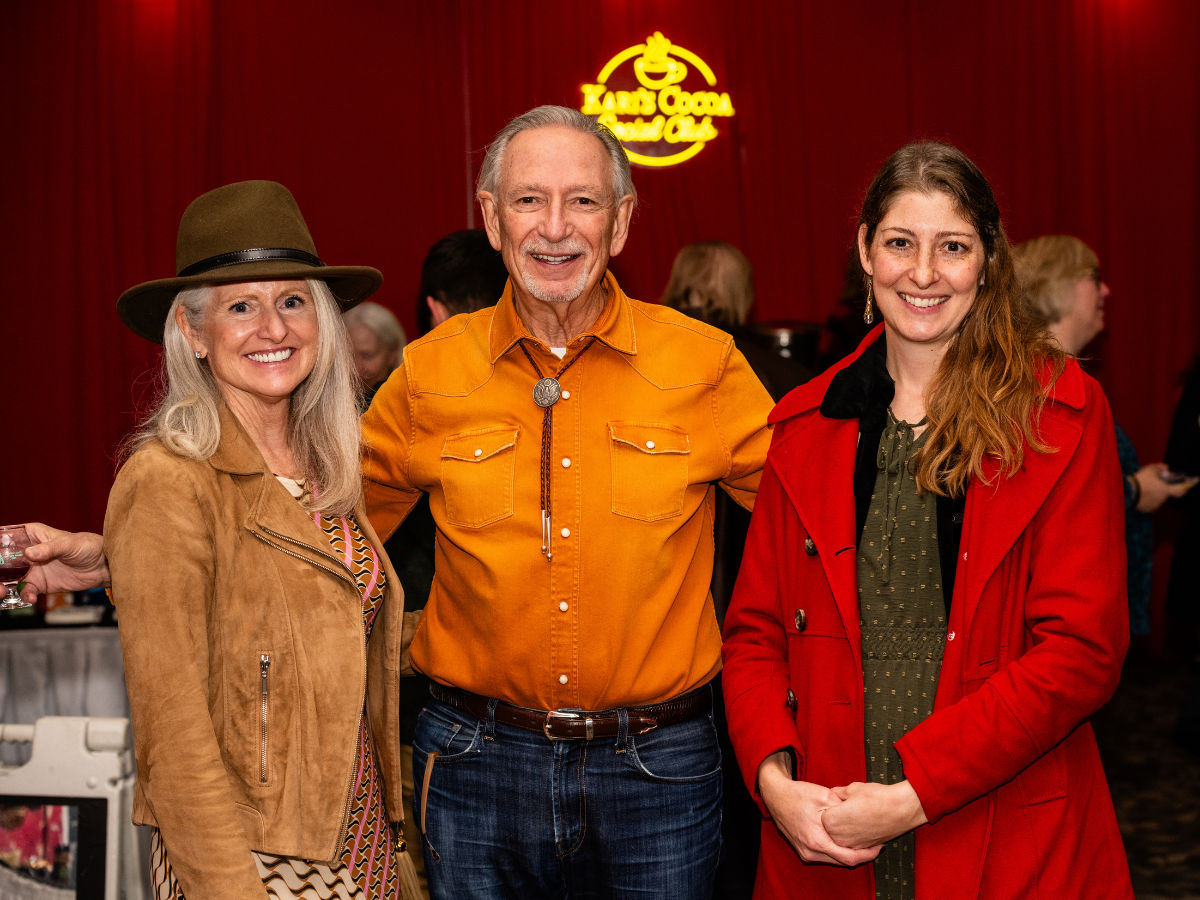 Three smiling people standing together at an indoor event, with a red background and a neon sign reading 'Texas Coop' in the background.