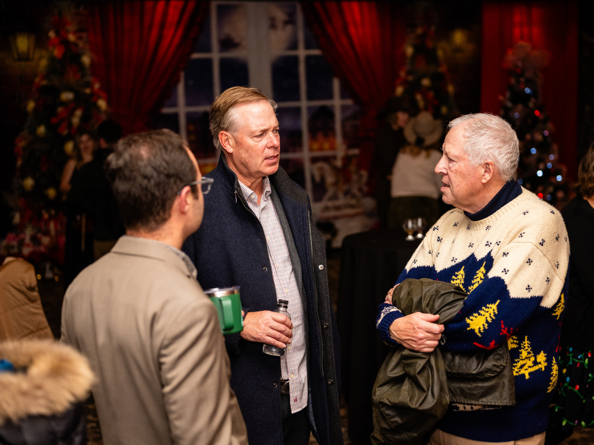 Three men having a conversation at a Christmas party, with Christmas trees, decorations, and red curtains in the background.