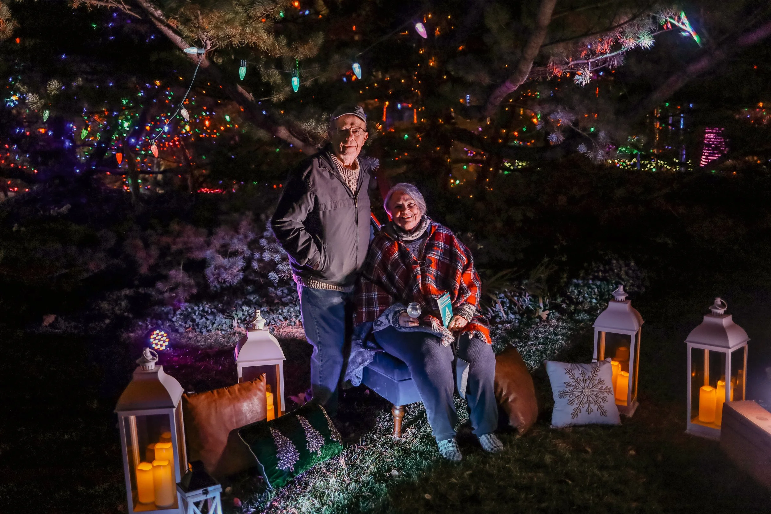 Two elderly women and a man outdoors at night during Christmas. One woman sits on a chair holding a drink, while the other stands beside her. They are surrounded by lit lanterns, pillows, and Christmas lights on trees.