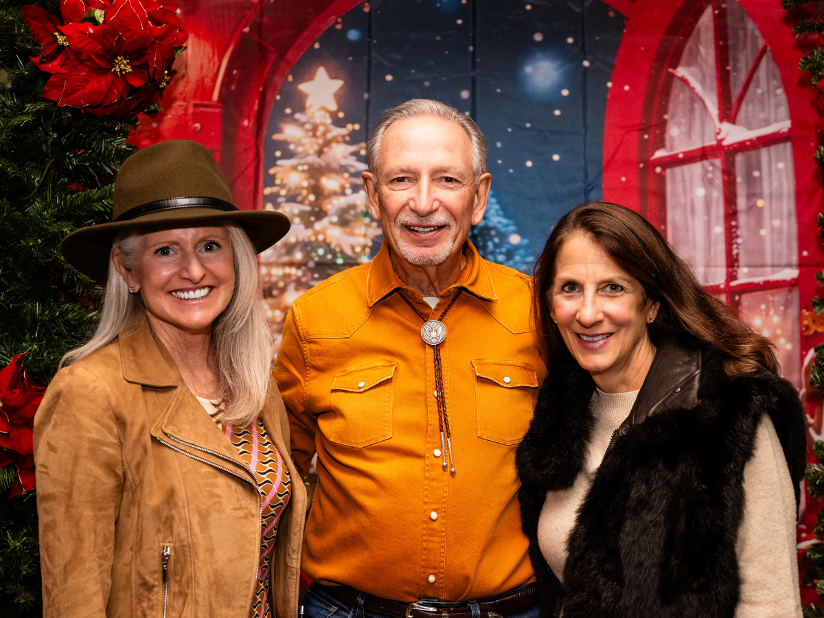 Three smiling people posing in front of a festive Christmas backdrop with a decorated tree and window scene, surrounded by holiday decorations.