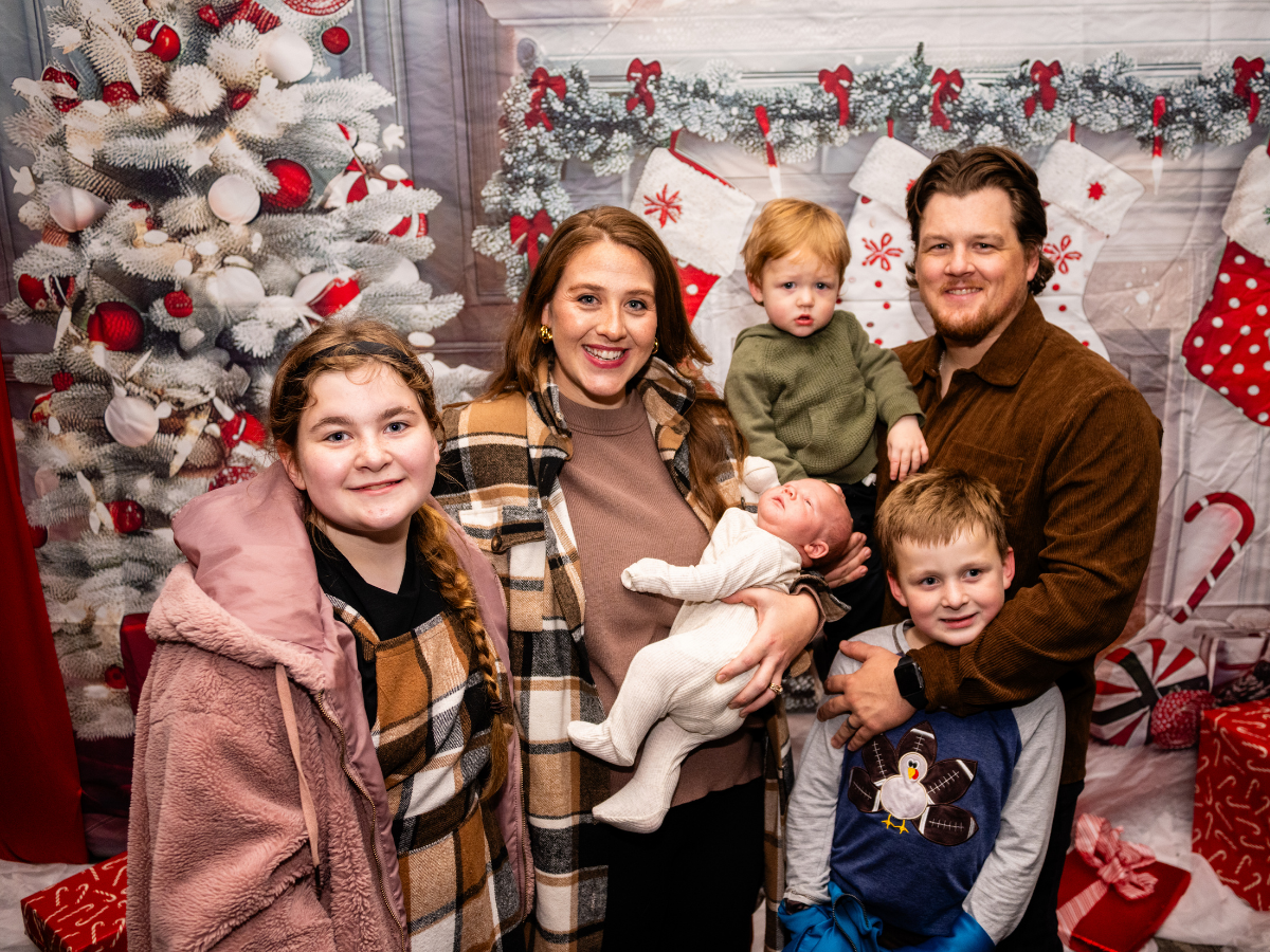A family of six celebrating Christmas, standing in front of a festive backdrop with a decorated white Christmas tree, stockings, and holiday decorations.