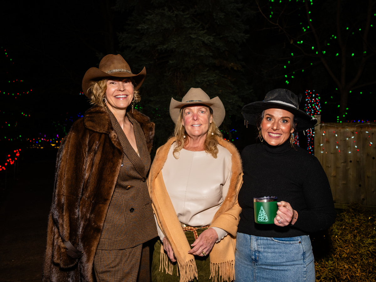 Three women in cowboy hats and fall clothing smiling at an outdoor nighttime event decorated with colorful Christmas lights.