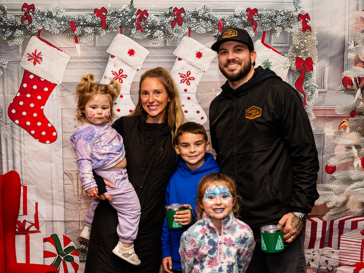 A family of five posing in front of a festive Christmas backdrop with stockings and holiday decorations. The children are smiling, with one girl painted as a face painter, and the others holding green mugs.