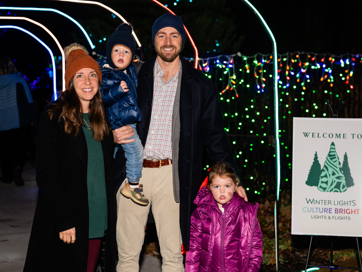 A family of four smiling at night in front of colorful Christmas lights, including a woman, a man, a young boy on the man's shoulder, and a young girl in a pink jacket.