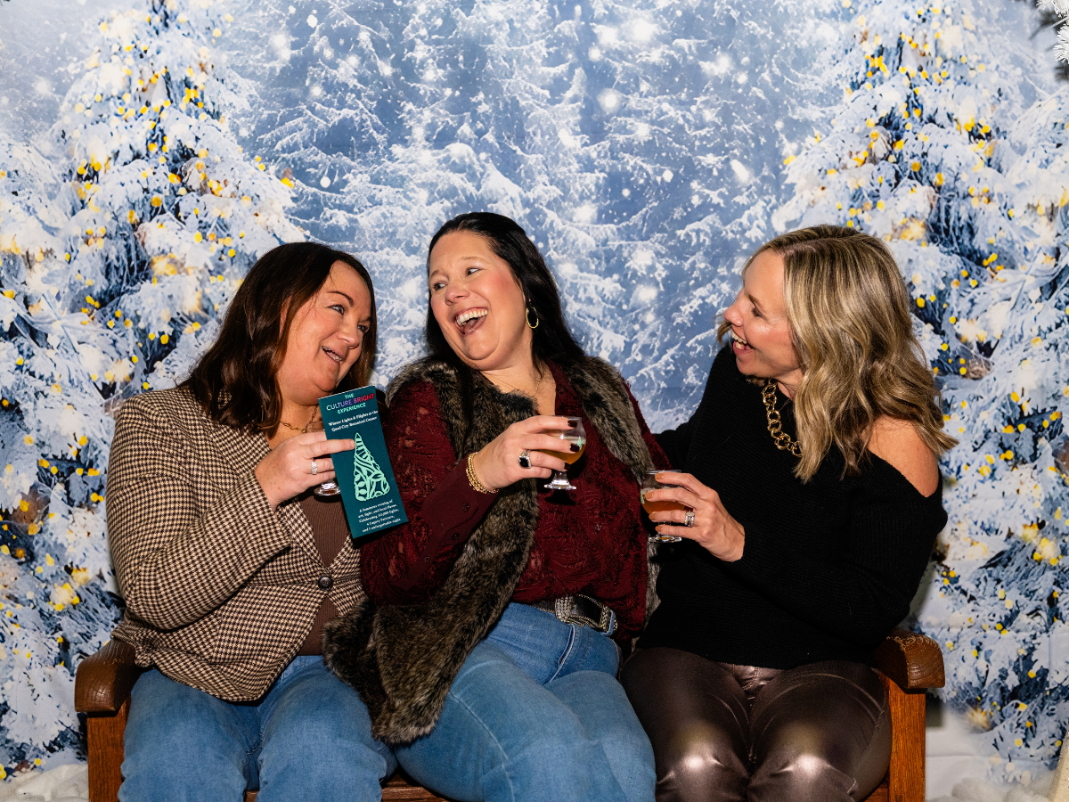 Three women sitting on a bench, laughing and clinking glasses in front of a winter snow-covered forest backdrop, with Christmas trees decorated with yellow ornaments.