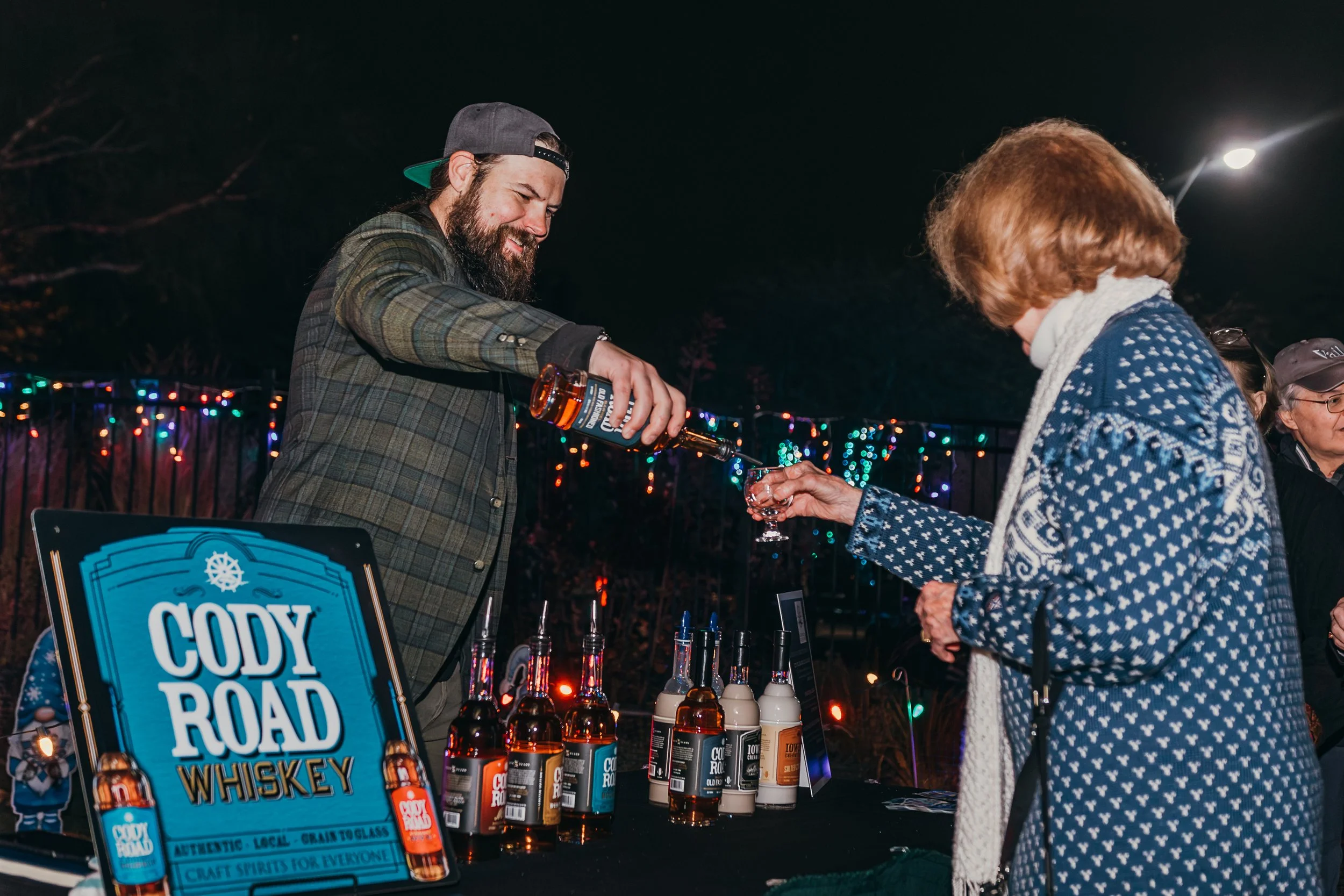 A man is pouring whiskey into a glass held by an older woman at an outdoor event with colorful string lights in the background. A table displays various bottles of Cody Road whiskey and a promotional sign.