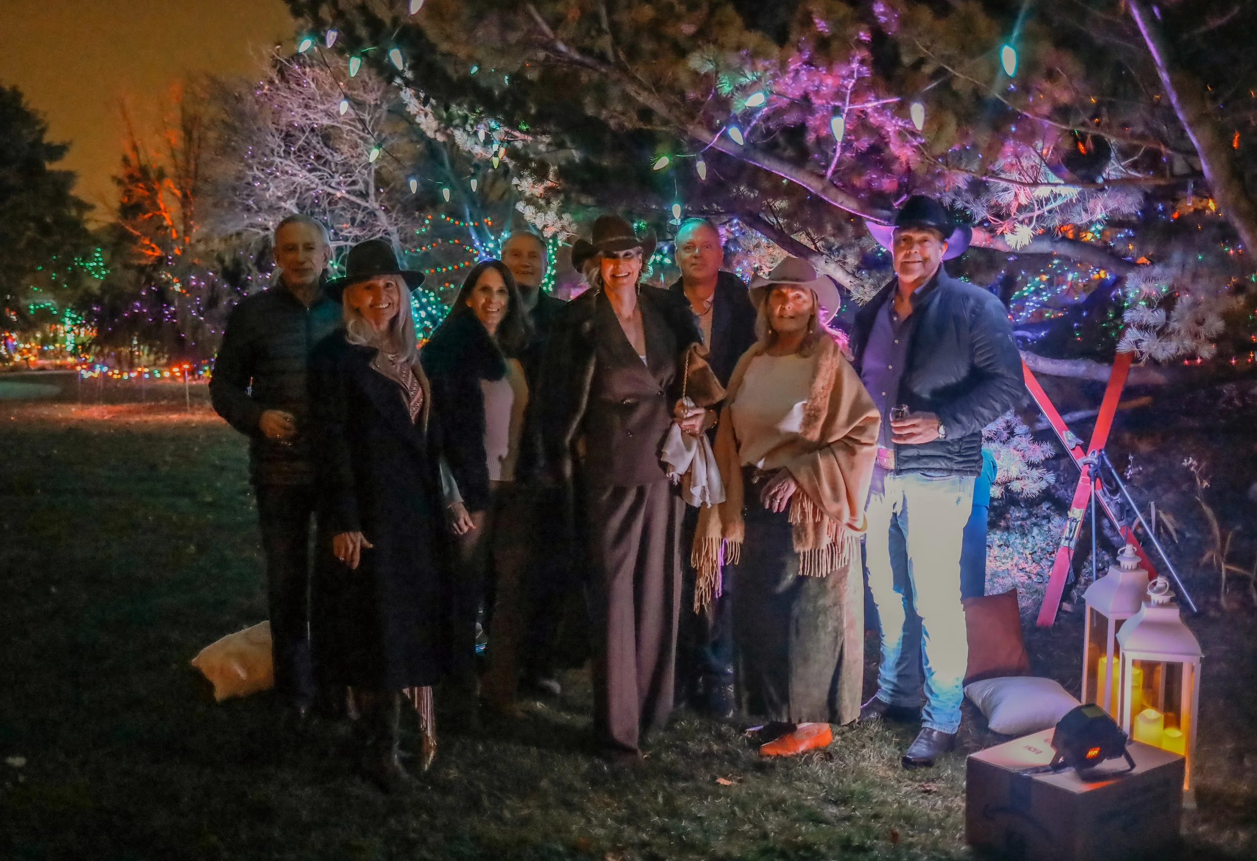 A group of nine people gathered outdoors at night, standing under a large tree decorated with colorful string lights. Some individuals are wearing cowboy hats, and a few are holding drinks. The scene is illuminated by festive lights and surrounded by