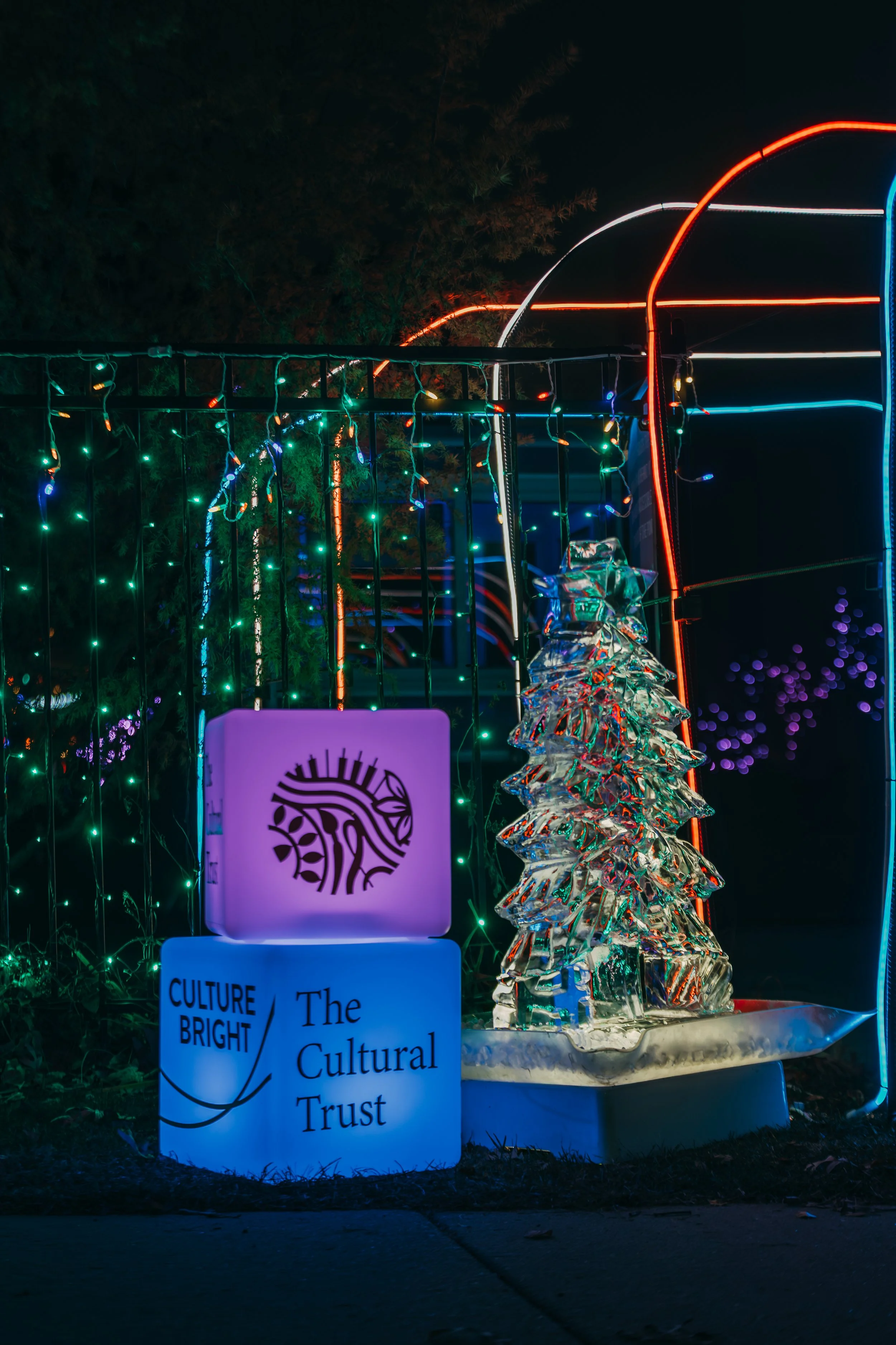 A decorative display featuring a glass sculpture of a Christmas tree, illuminated in vibrant colors, next to two illuminated signs that read "CULTURE BRIGHT" and "The Cultural Trust," with colorful string lights and neon accents in the background.