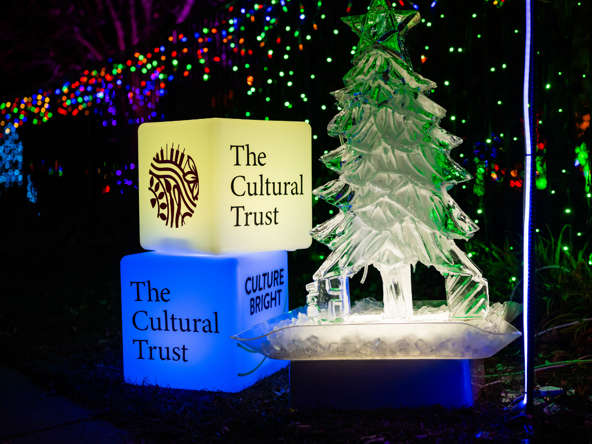 Colorfully illuminated display with glowing blocks reading "The Cultural Trust" and "CULTURE BRIGHT" next to a clear ice sculpture of a Christmas tree, decorated with colorful lights in the background.