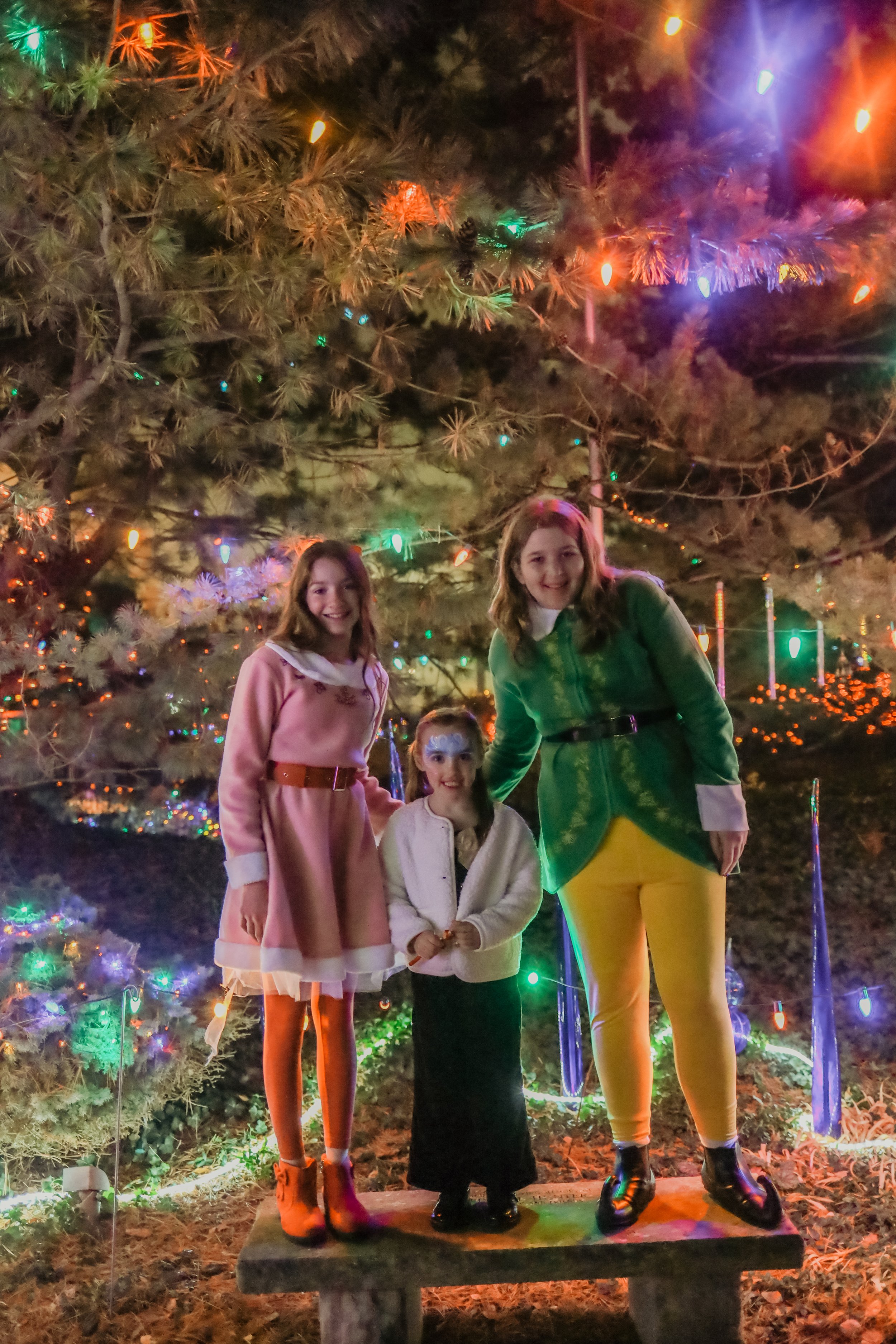 Three girls standing on a wooden bench in front of a decorated Christmas tree with colorful lights at night.