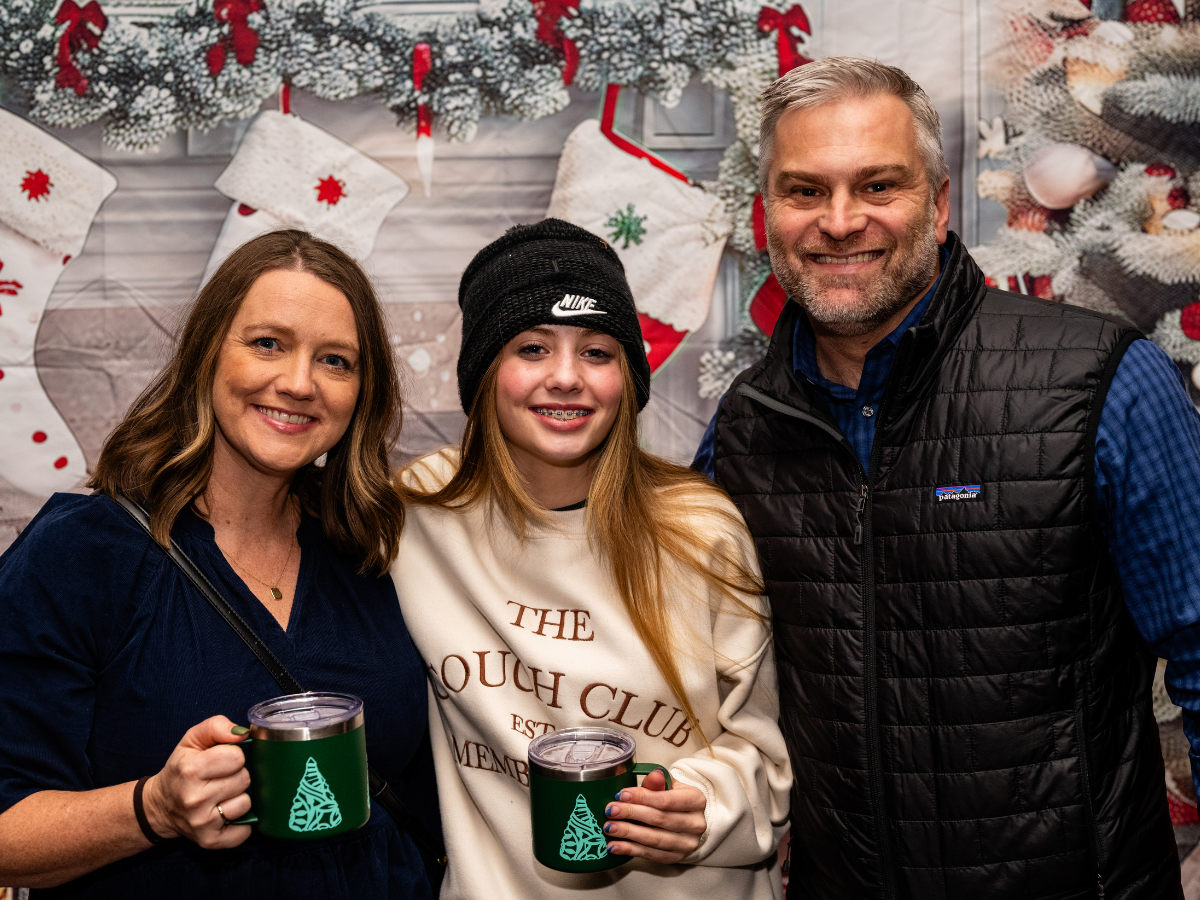 Three people smiling and holding holiday-themed mugs, standing in front of Christmas stockings and decorations.