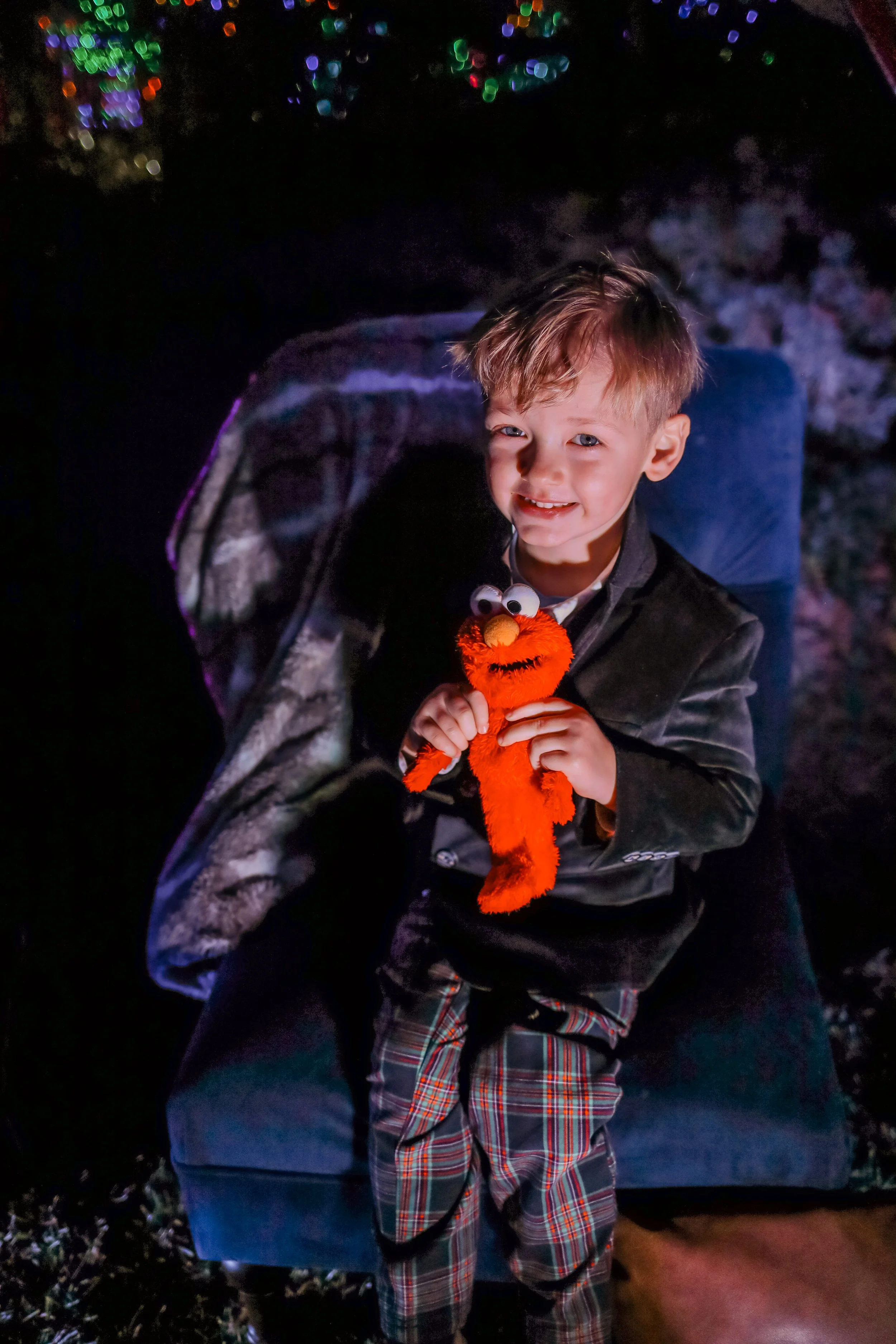 A young boy with light-colored hair and blue eyes sitting on a blue chair outdoors at night, holding a plush Elmo toy with googly eyes and an orange nose, smiling at the camera.